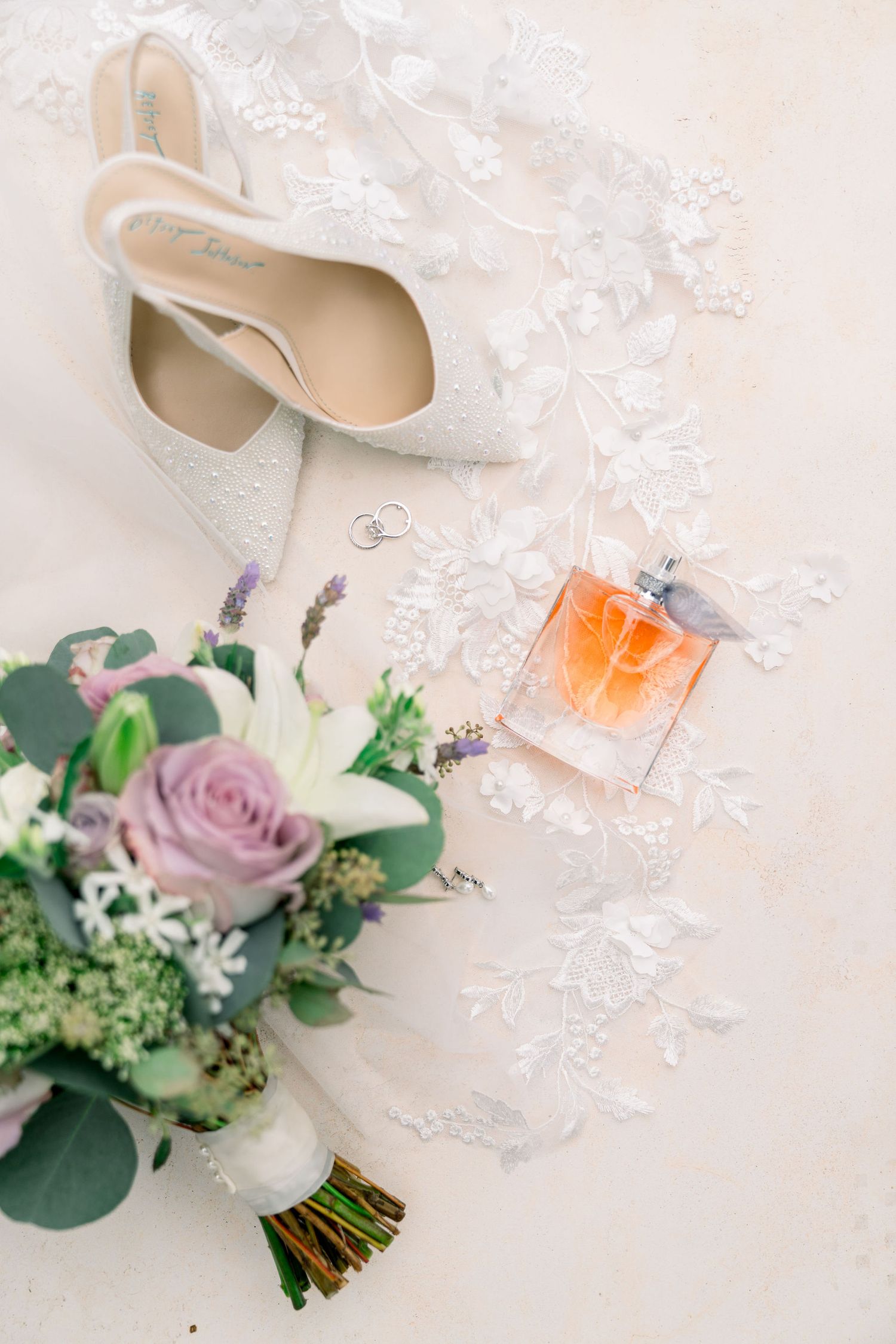 Elegant wedding details with white heels, perfume bottle, and pink rose bouquet arranged on delicate lace.