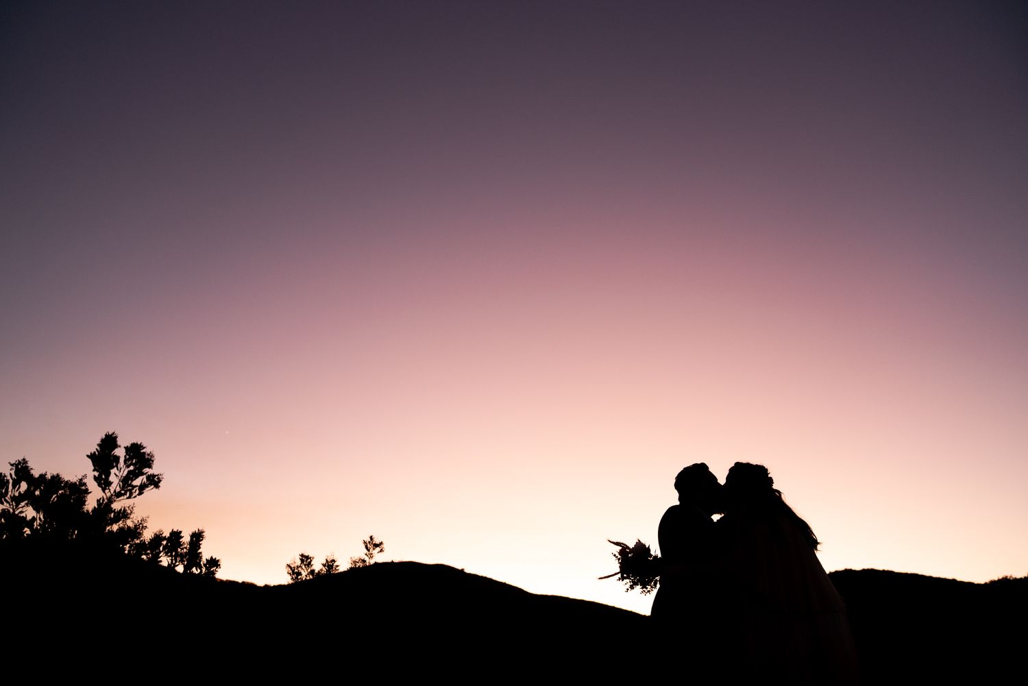 Silhouetted couple sharing an intimate moment against a dreamy purple sunset sky with mountain landscape backdrop.