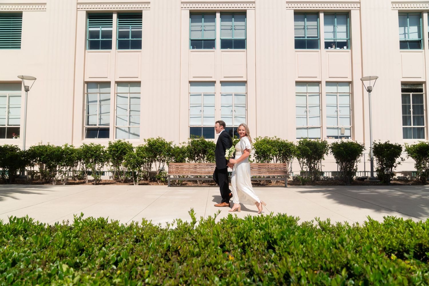 A romantic couple embraces in front of a grand white building with tall windows and manicured hedges.