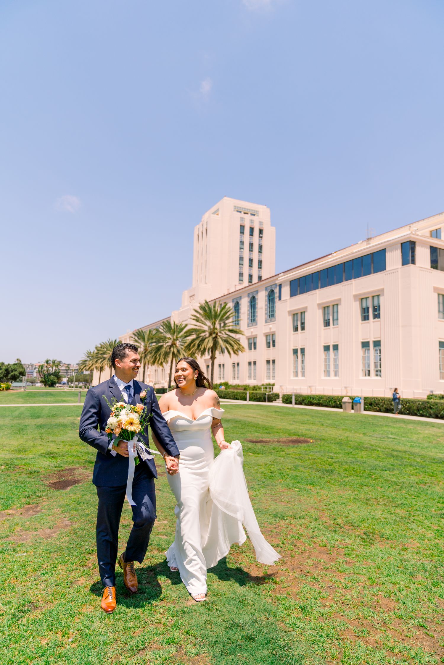Two people in wedding attire playfully dance together on a green lawn in front of a white building with palm trees.