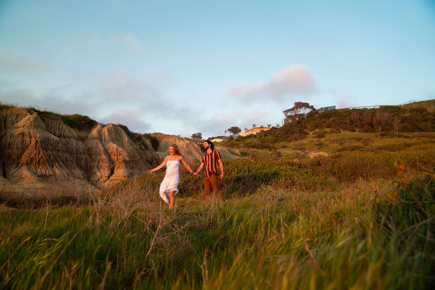 Two people run joyfully through tall grass on a coastal hillside with cliffs in the background.