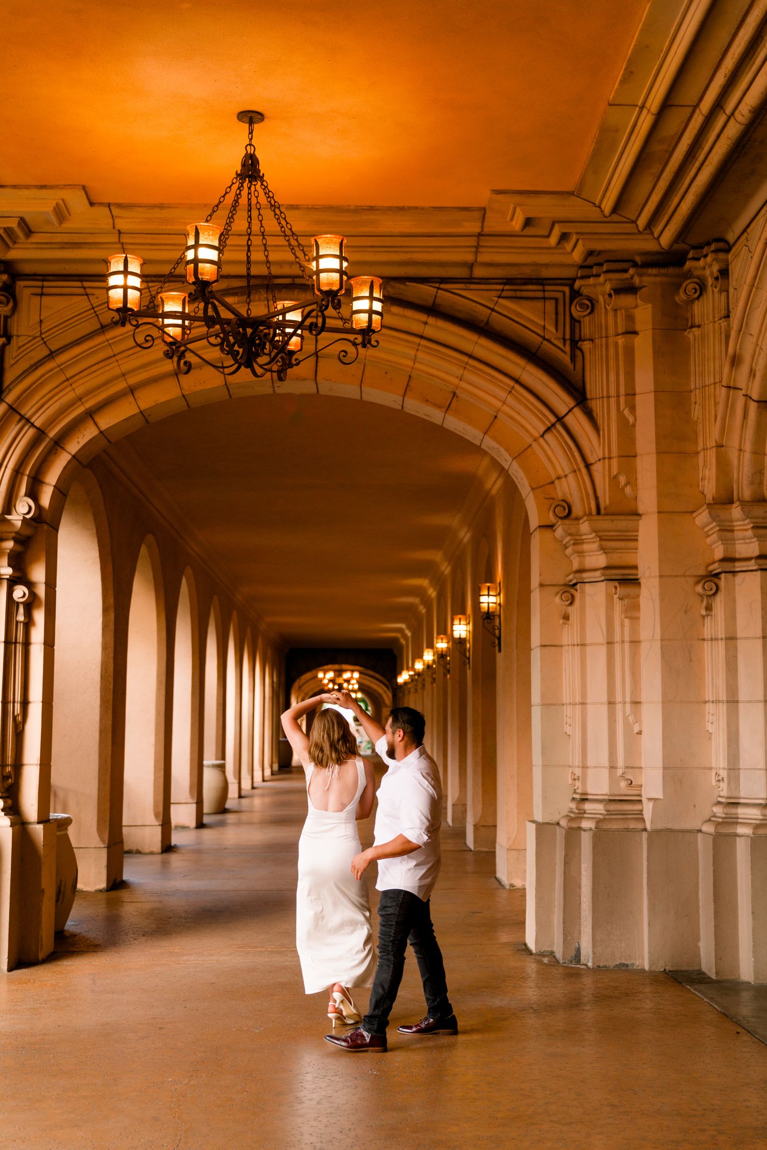 A couple dances together in an elegant arched colonnade hallway illuminated by warm hanging lanterns at sunset.
