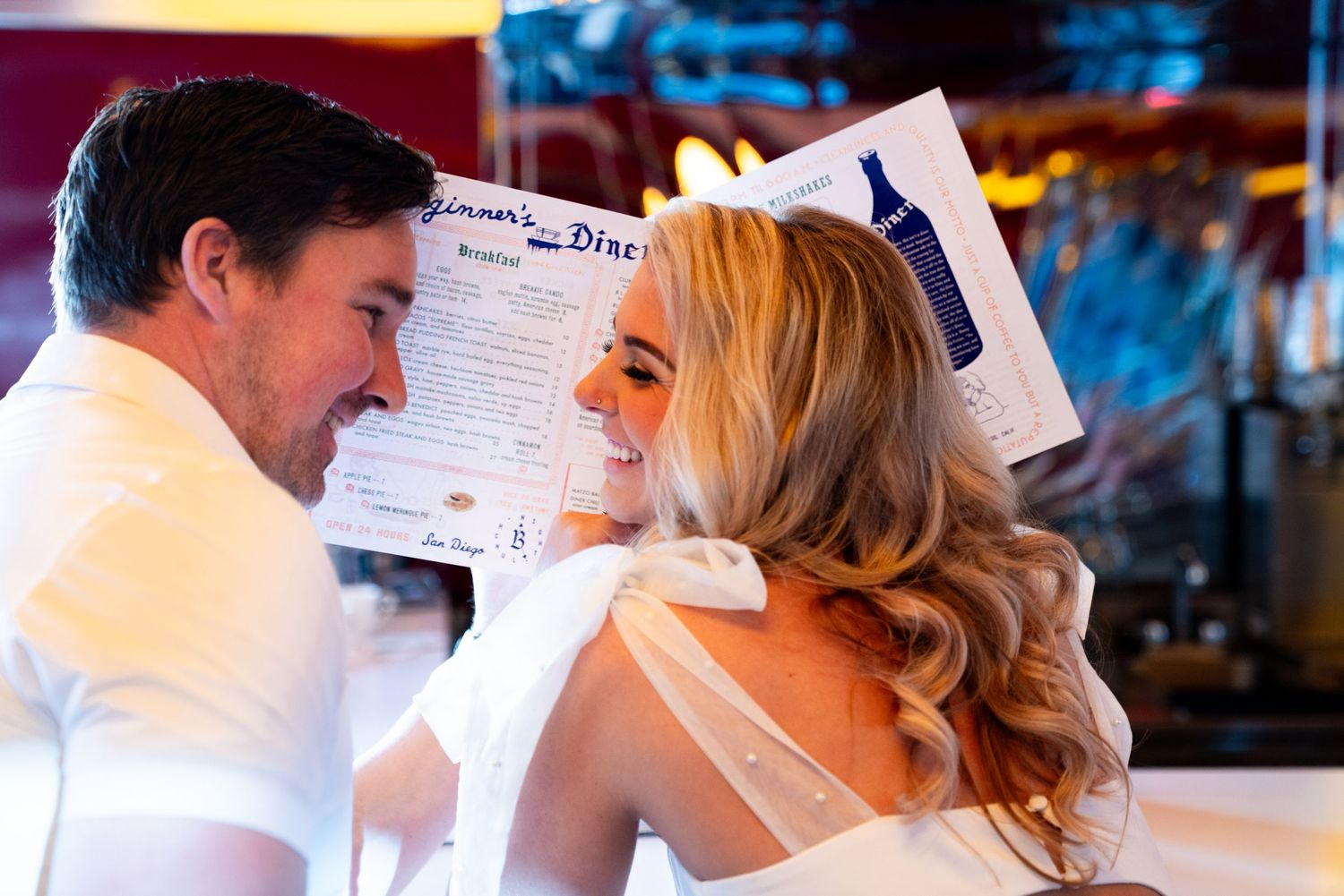 A couple at a diner shares a menu and laughs together, with a white bow and retro style decor in view.