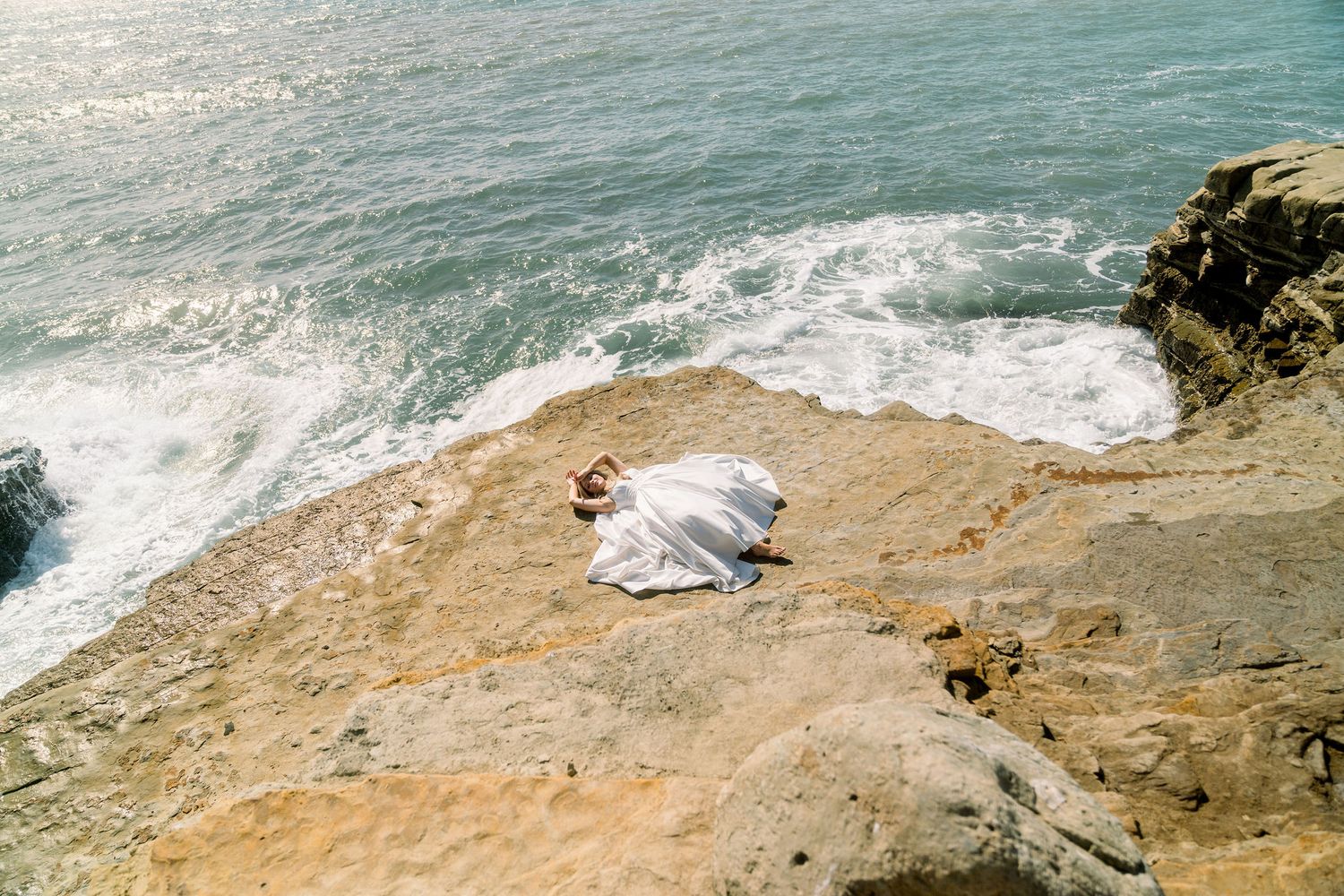 A white blanket spread out on a rocky coastal cliff overlooking turquoise ocean waves crashing below.