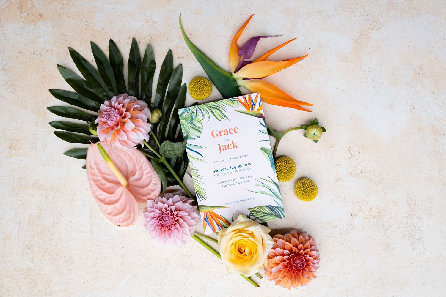 Tropical floral arrangement with bird of paradise, dahlias, and palm leaves surrounding a wedding invitation card.