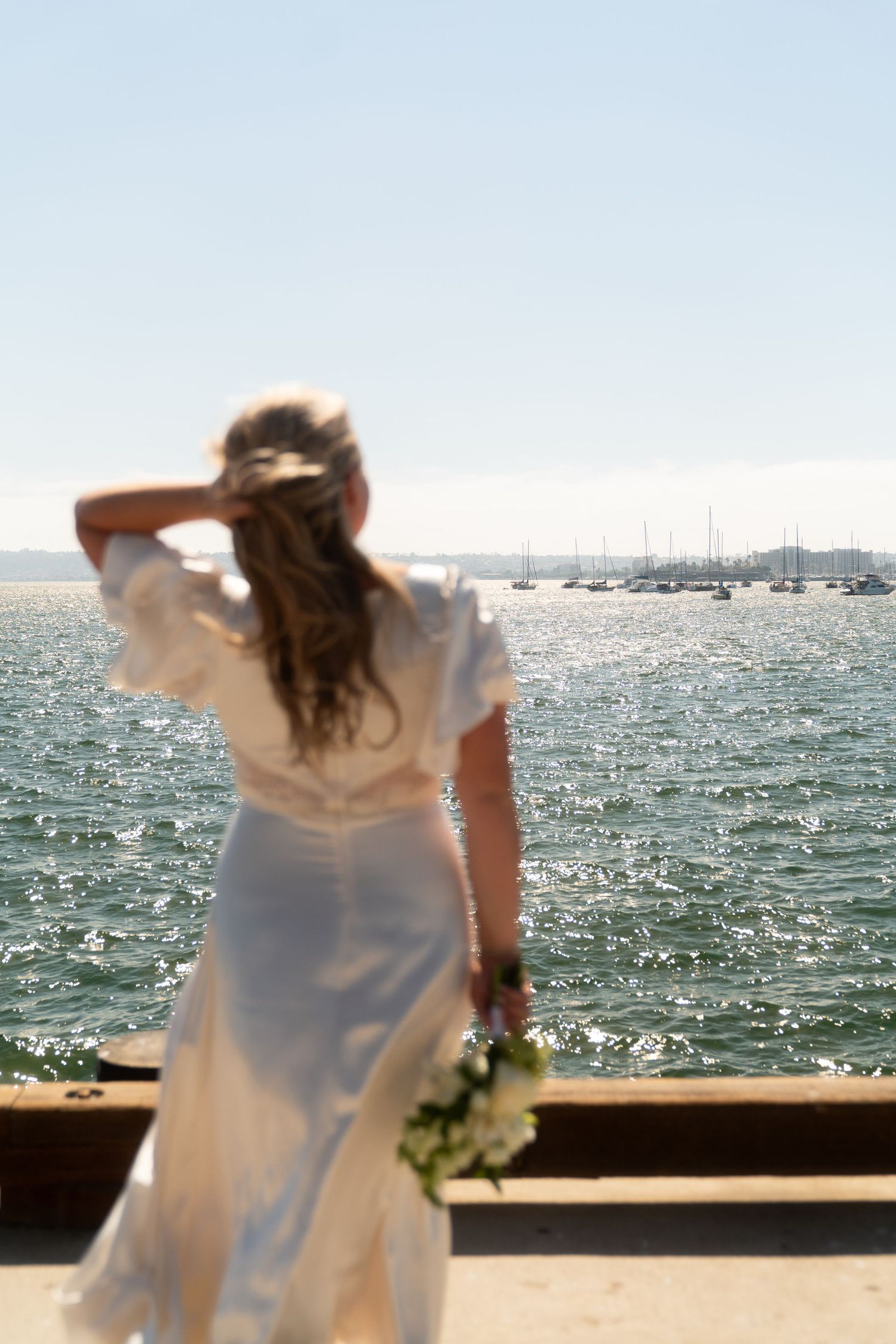 A figure in a flowing white dress stands on a waterfront deck overlooking a sparkling ocean on a bright sunny day.