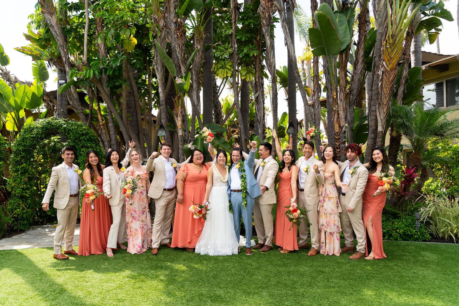 Wedding party in colorful coral and beige dresses poses in a lush tropical garden setting for group photos.