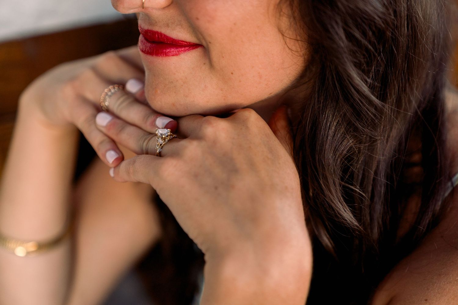 Close up detail shot showing red lipstick, dark wavy hair, and a sparkling engagement ring on a hand.