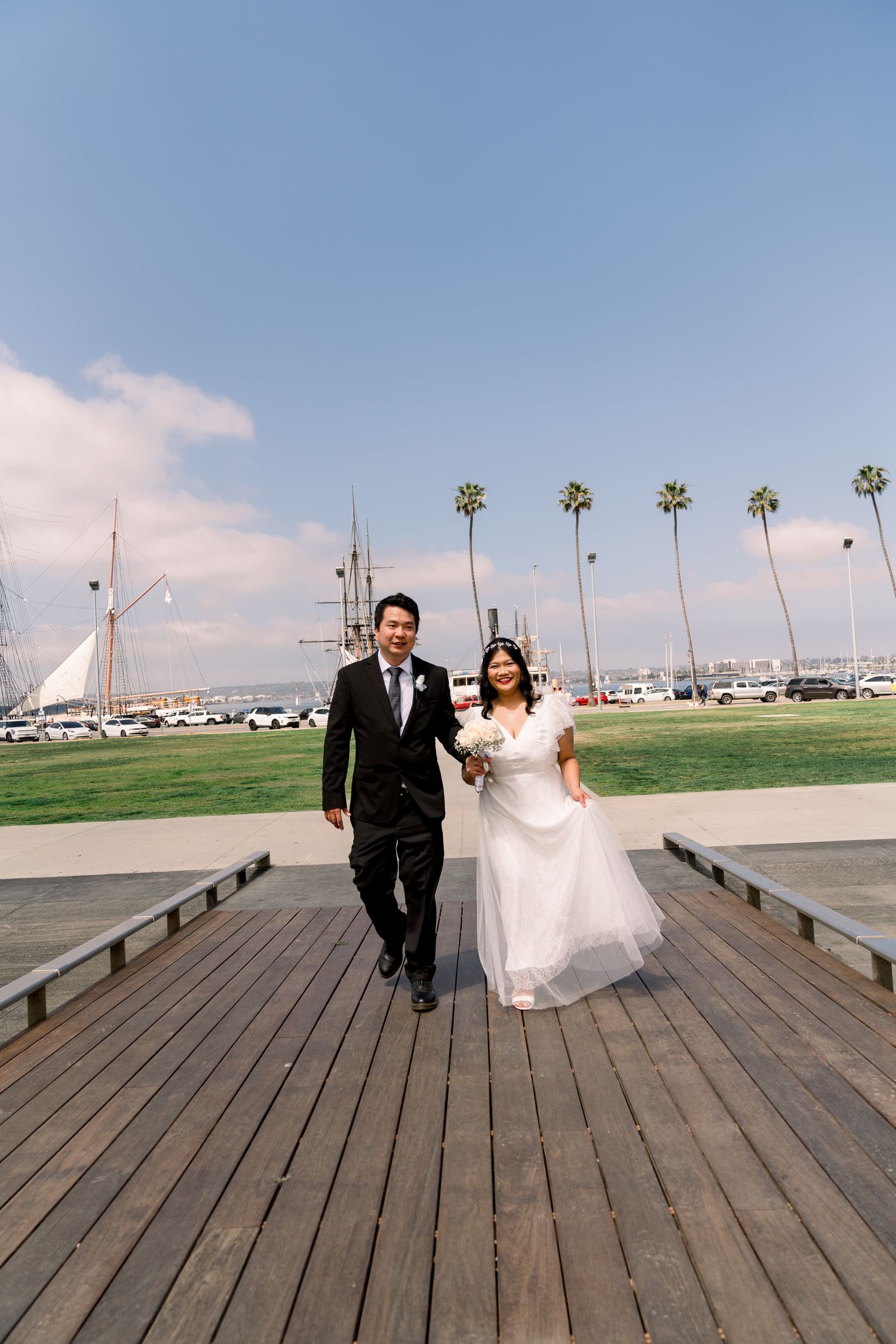 A wedding couple walks together on a wooden pier with palm trees and boats in the background.