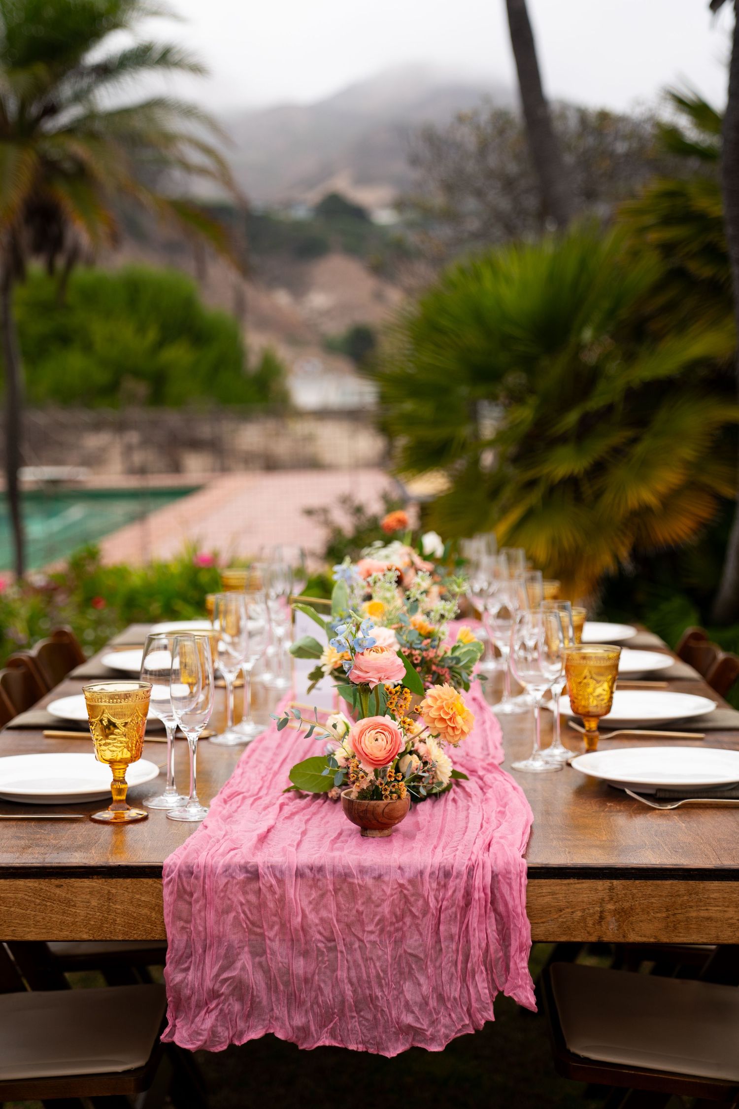 Elegant outdoor dining table with pink chiffon runner and floral centerpiece overlooking palm trees and mountains.