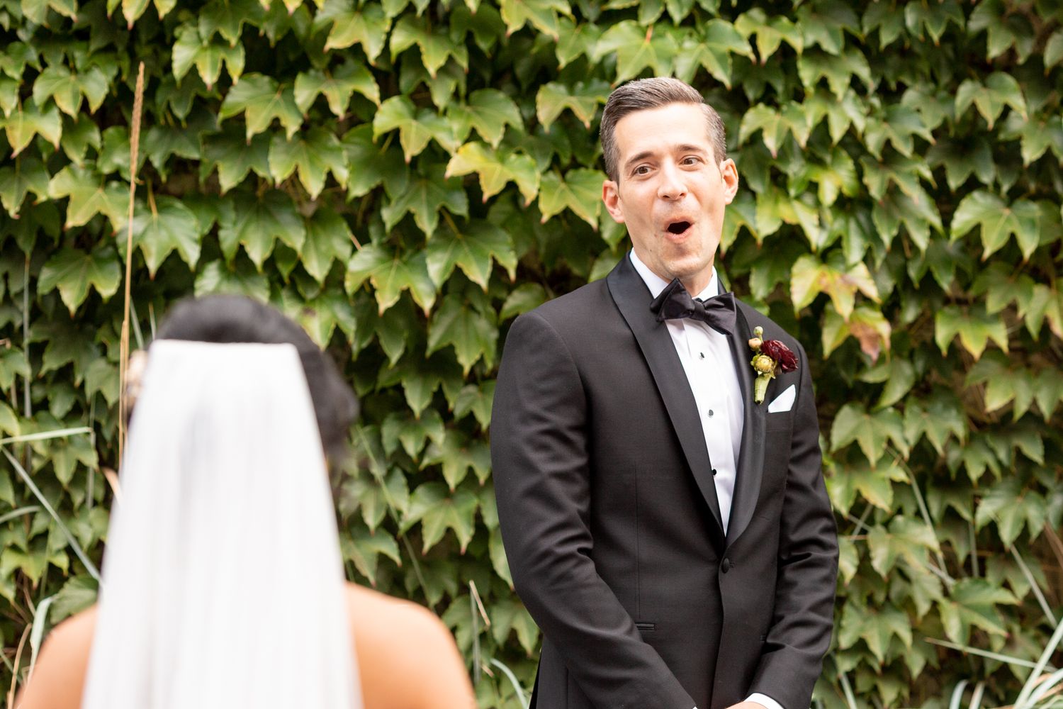 A groom in a black tuxedo and bowtie reacts with joy against an ivy-covered wall outside.