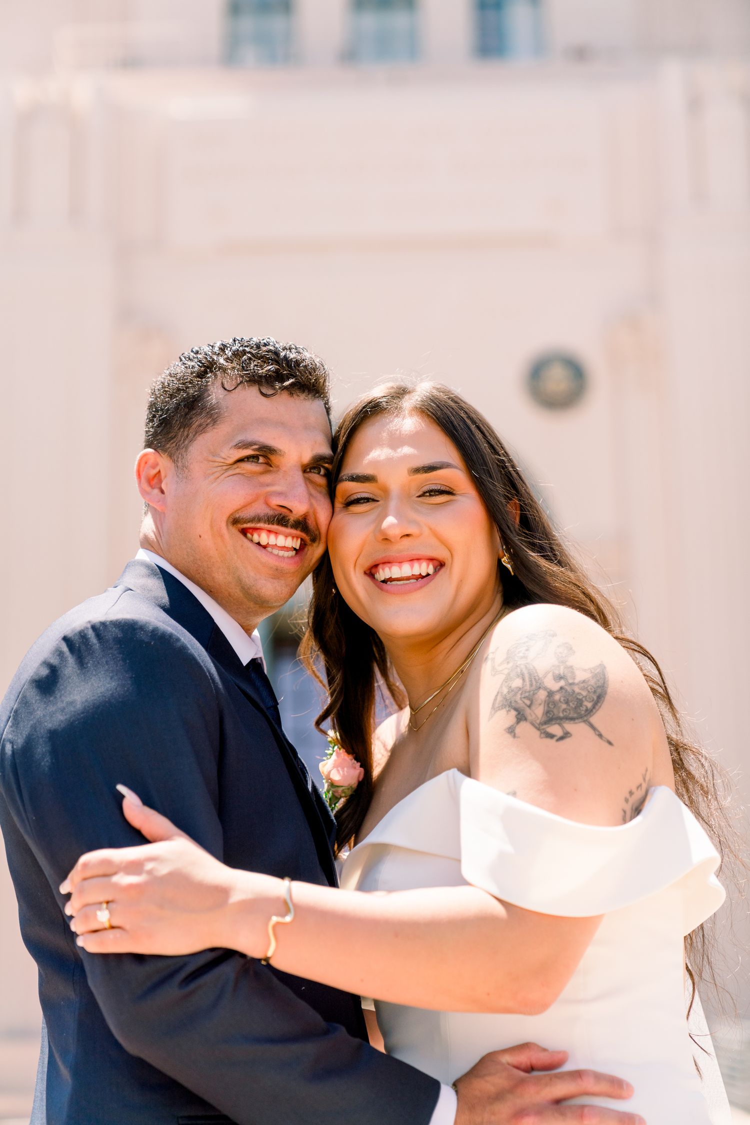 A joyful couple embraces and laughs together on their wedding day, with the bride wearing a white dress showing her arm tattoo.