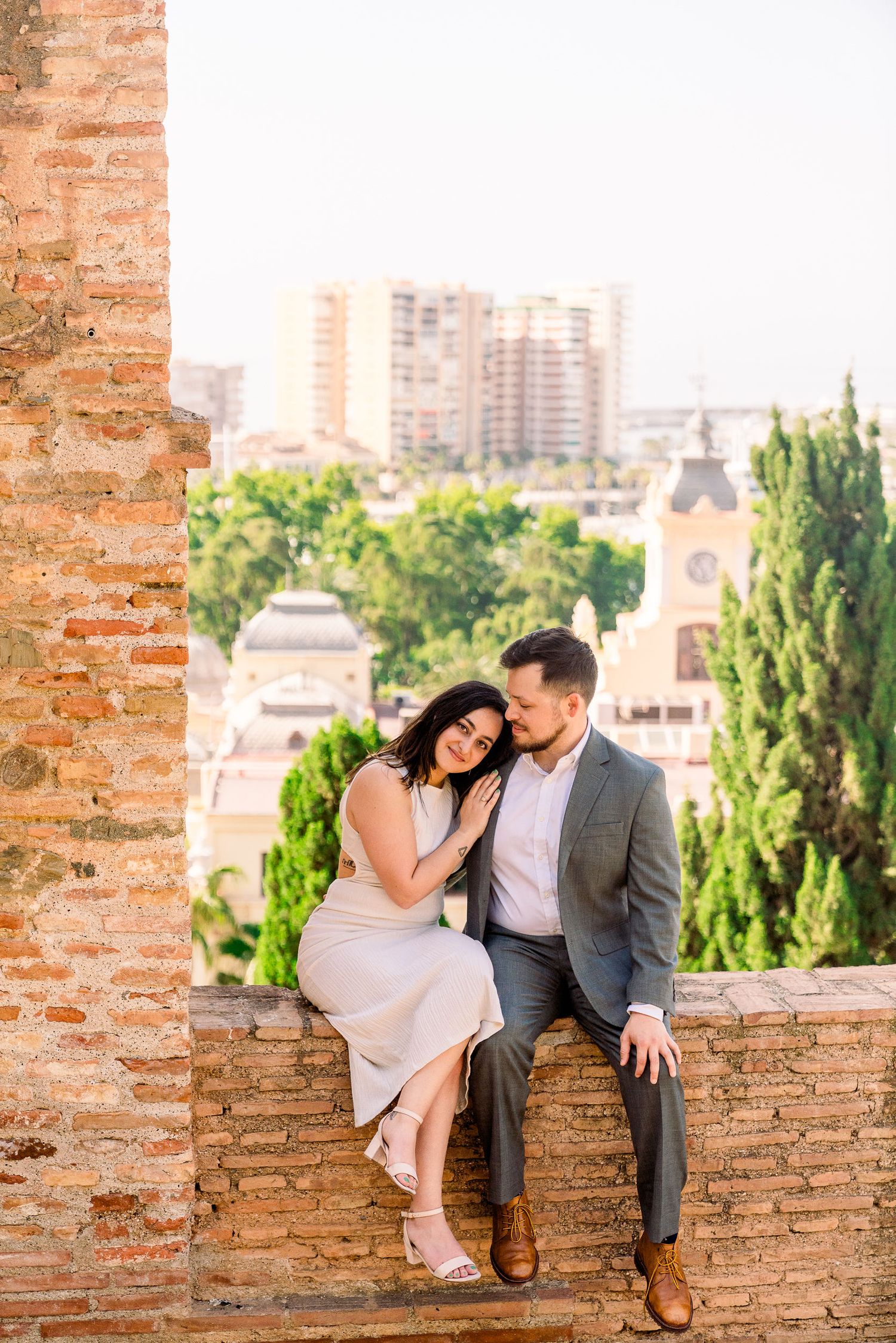 A romantic couple sits together on an ancient brick wall overlooking a European cityscape with trees and buildings in view.
