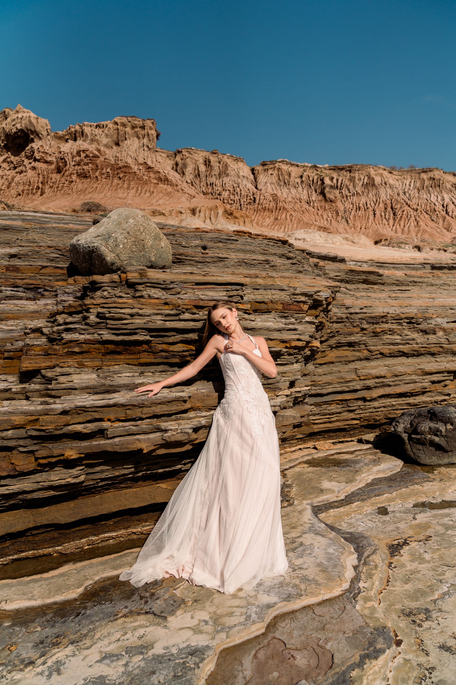 A figure in a flowing white wedding dress poses gracefully on rocky coastal formations against dramatic red cliffs and blue sky.