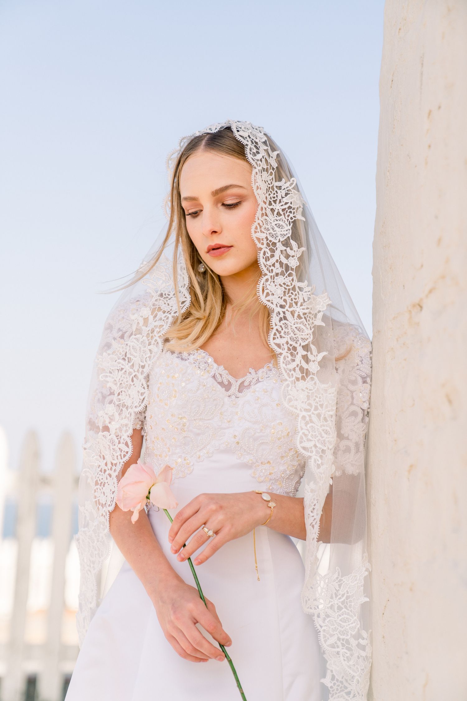A figure in a romantic white bridal gown and delicate lace veil poses elegantly against a light backdrop.