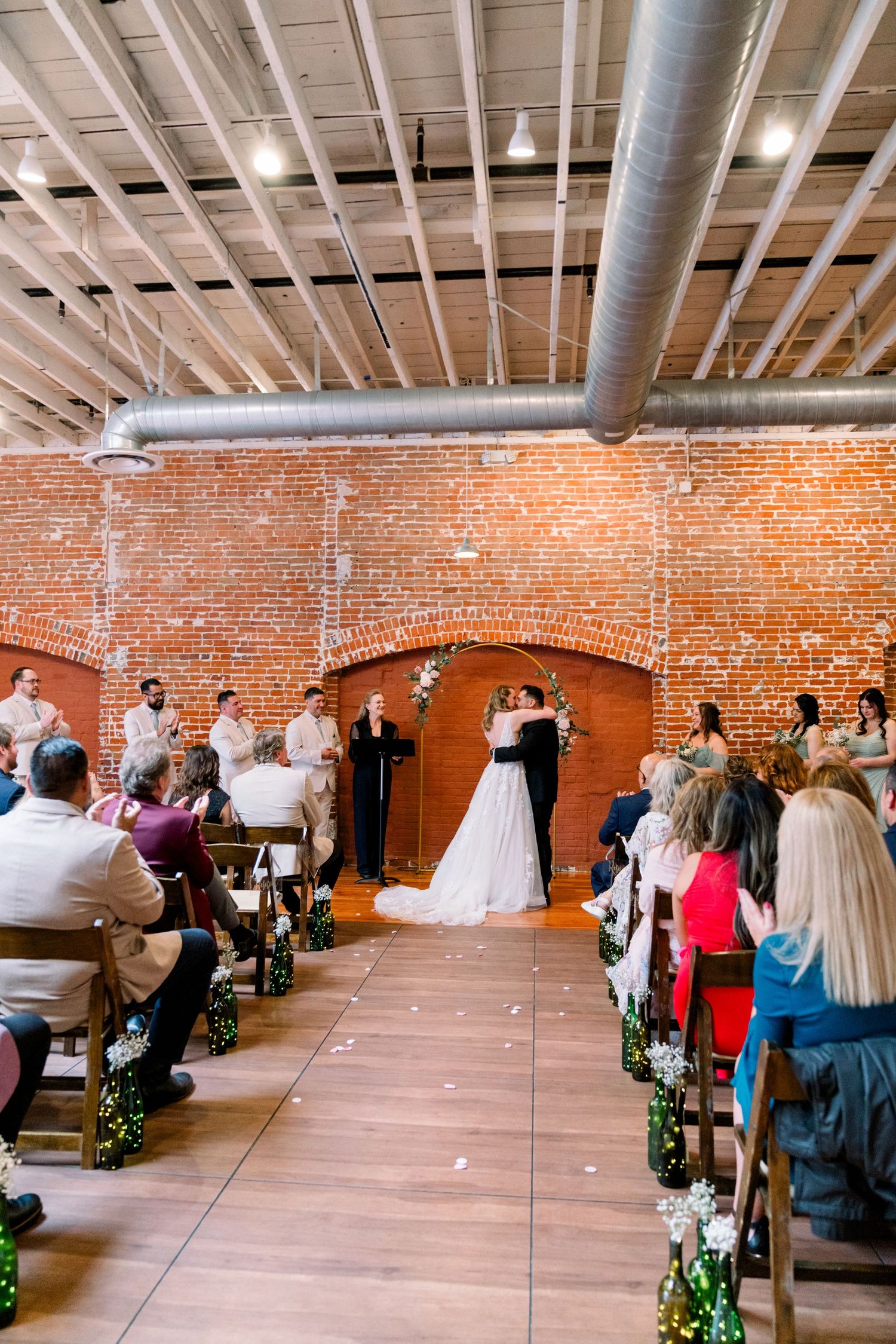 Indoor wedding ceremony in rustic industrial venue with exposed brick walls, wooden benches and overhead ductwork.