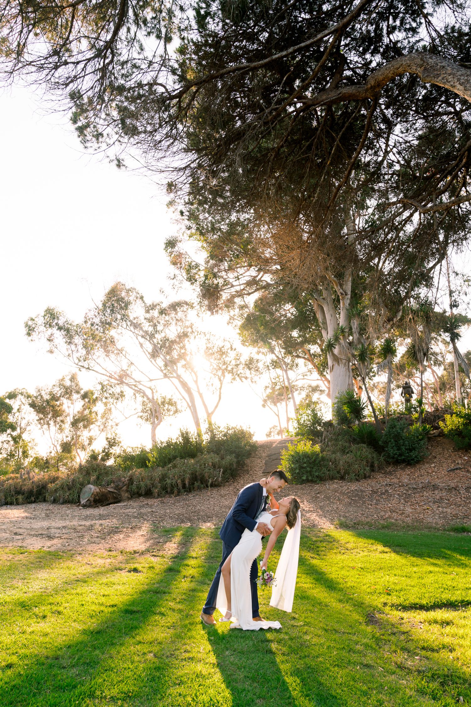 A couple shares an intimate moment on a grassy hillside at sunset with eucalyptus trees in the background.