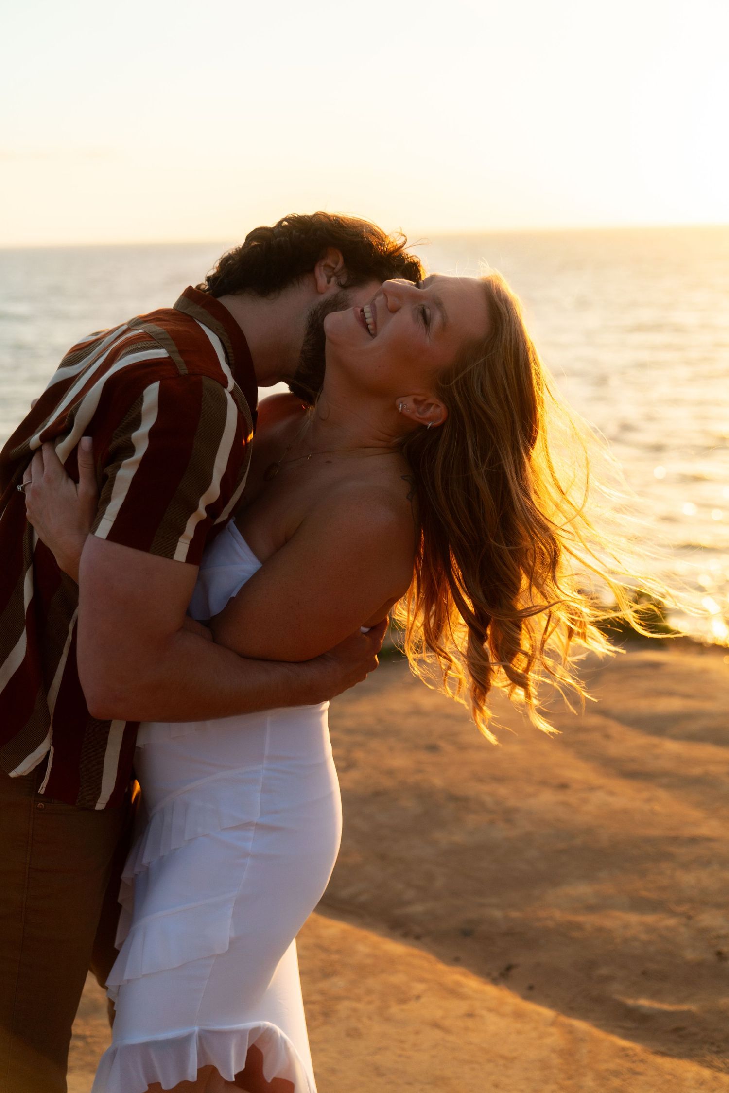 A romantic couple embraces on the beach at sunset with golden sunlight illuminating their intimate moment.