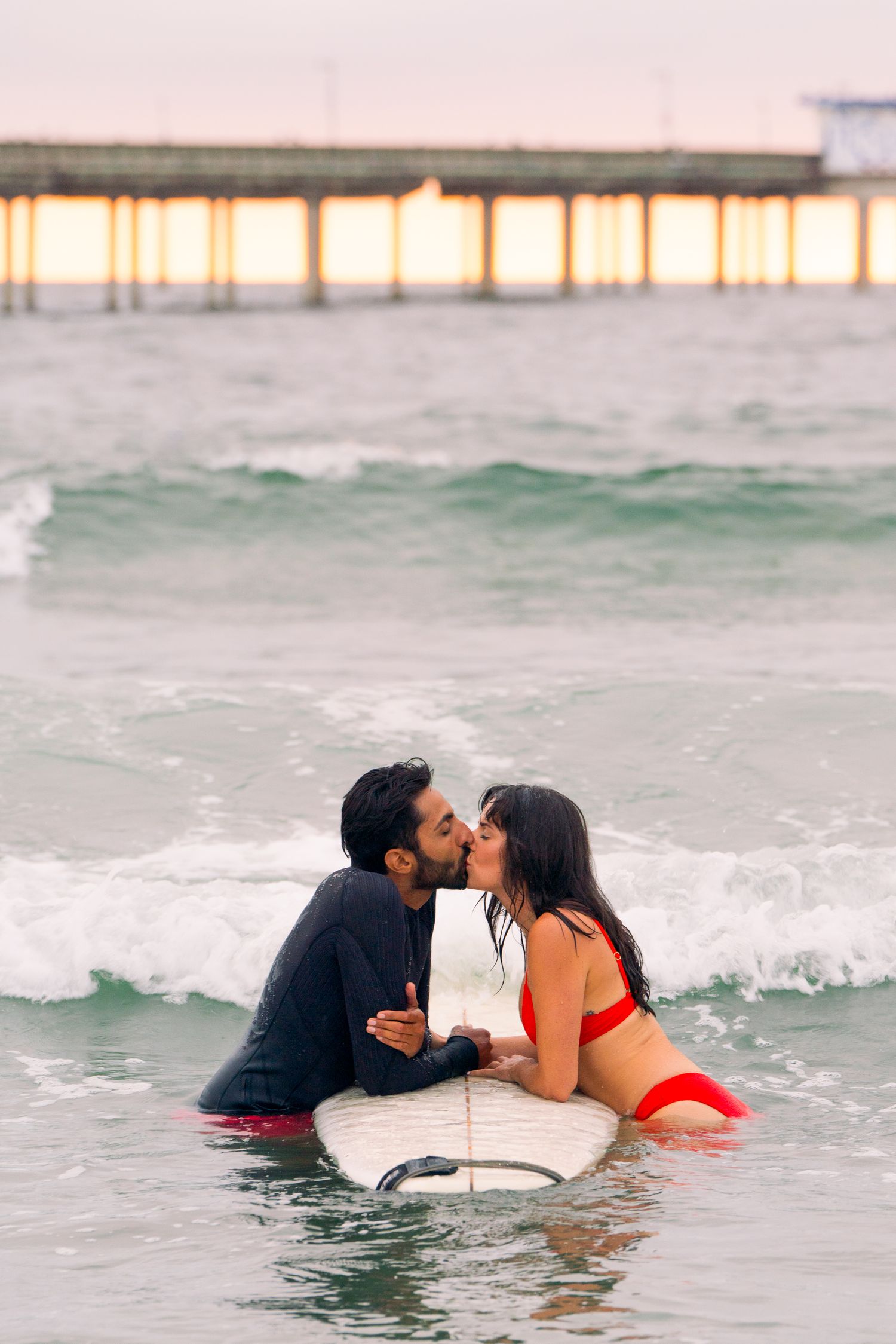 Two people share a kiss while lying on a surfboard in ocean waves at sunset with a pier visible in the background.