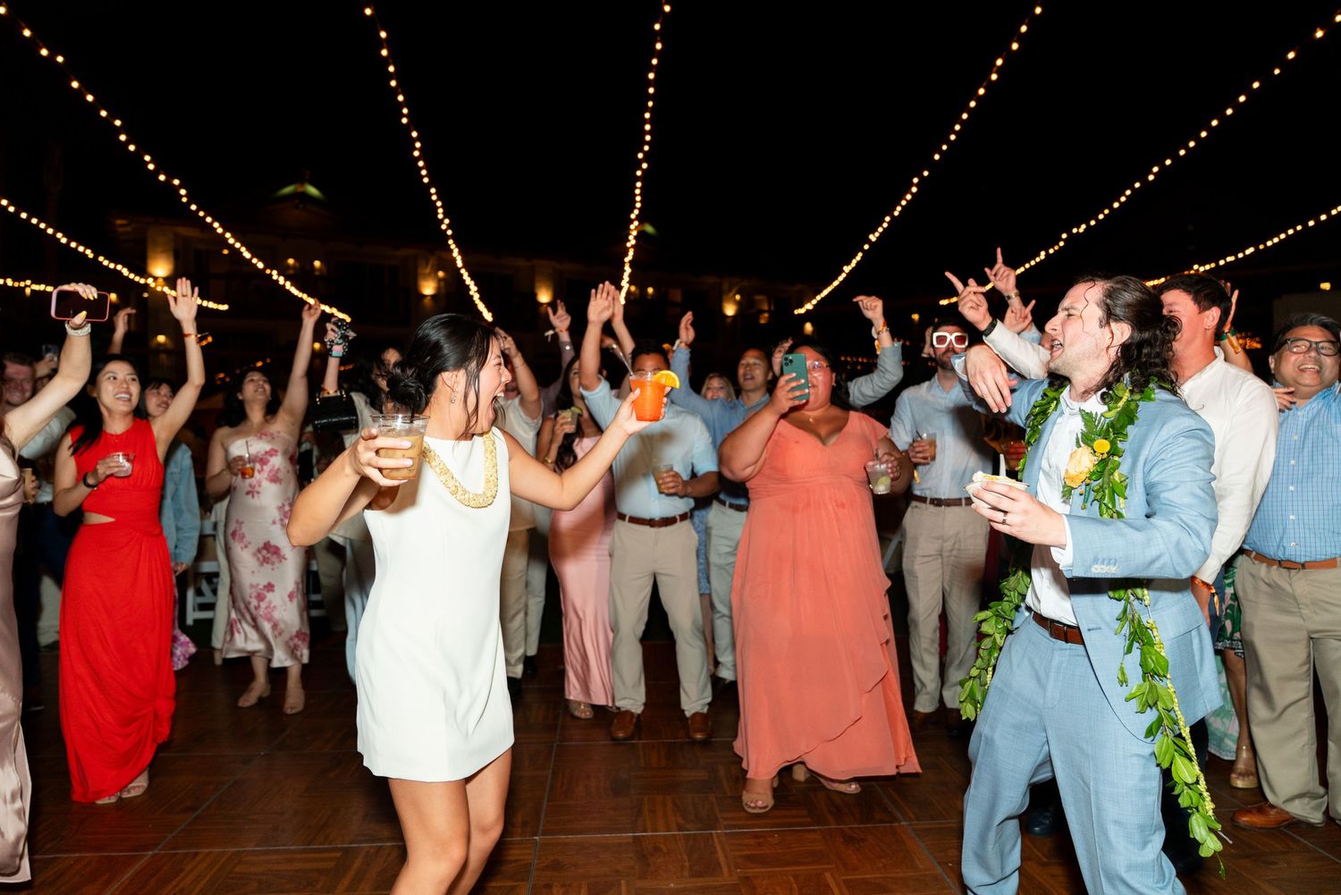 A lively group dances together under stringed lights at a festive outdoor reception party.