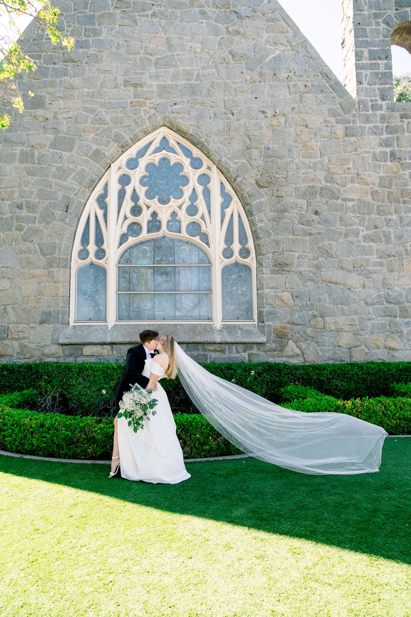 A flowing white veil cascades across lush green grass against a stone church wall with Gothic architectural details.