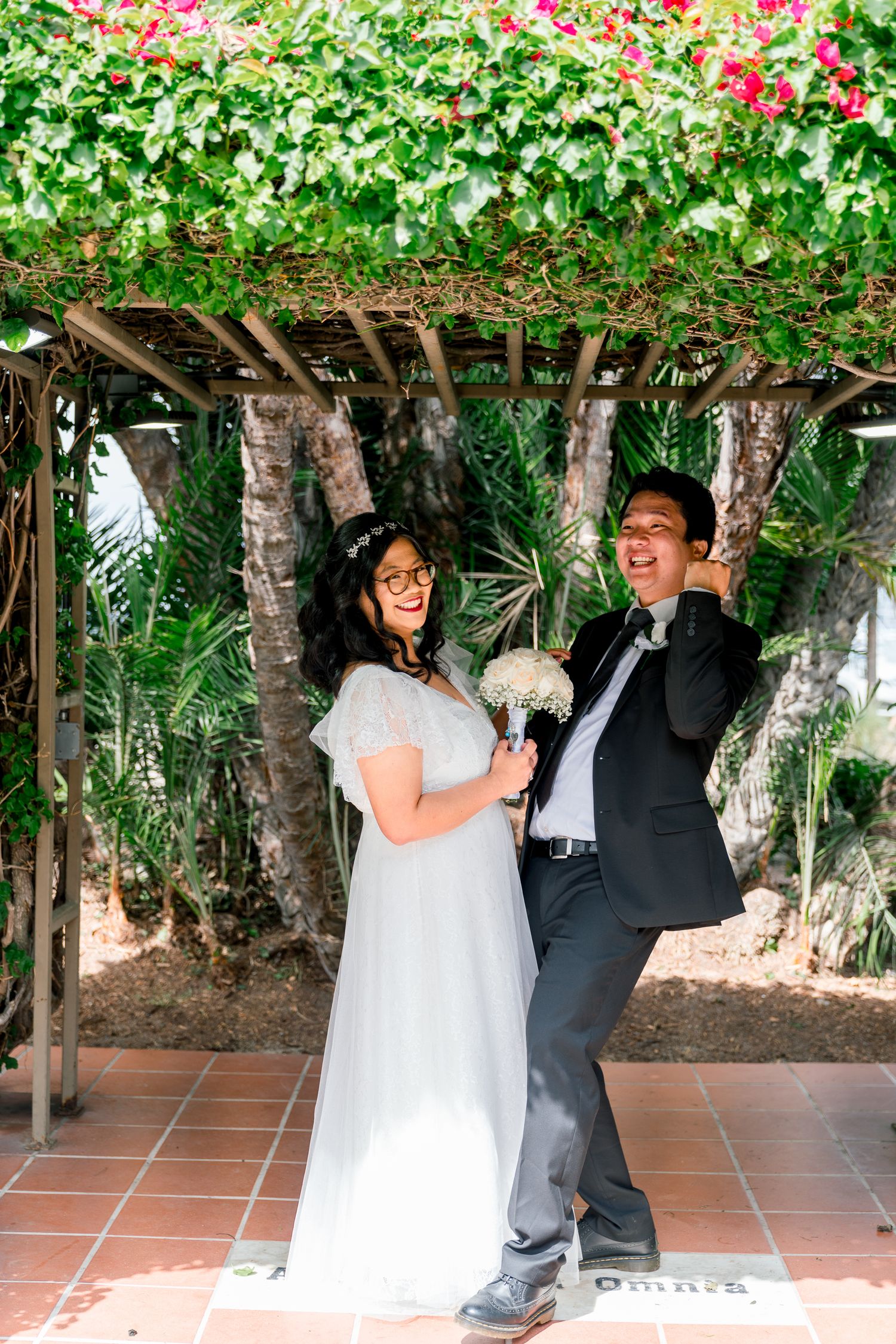 A happy couple shares a lighthearted moment under a floral pergola on their wedding day, surrounded by pink blooms and greenery.
