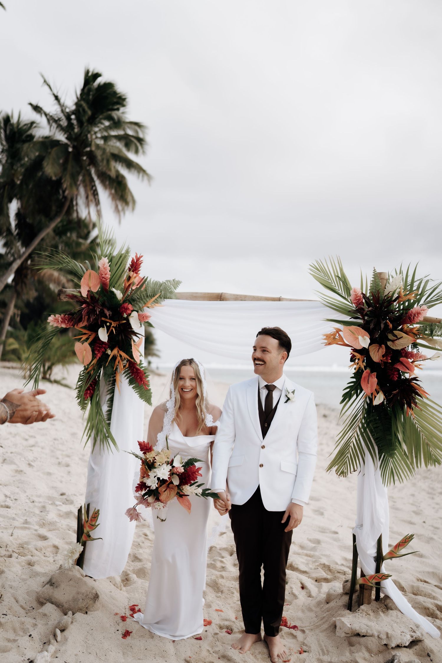 Bride and groom stand barefoot on sandy beach under tropical floral arch with palm trees and ocean backdrop.