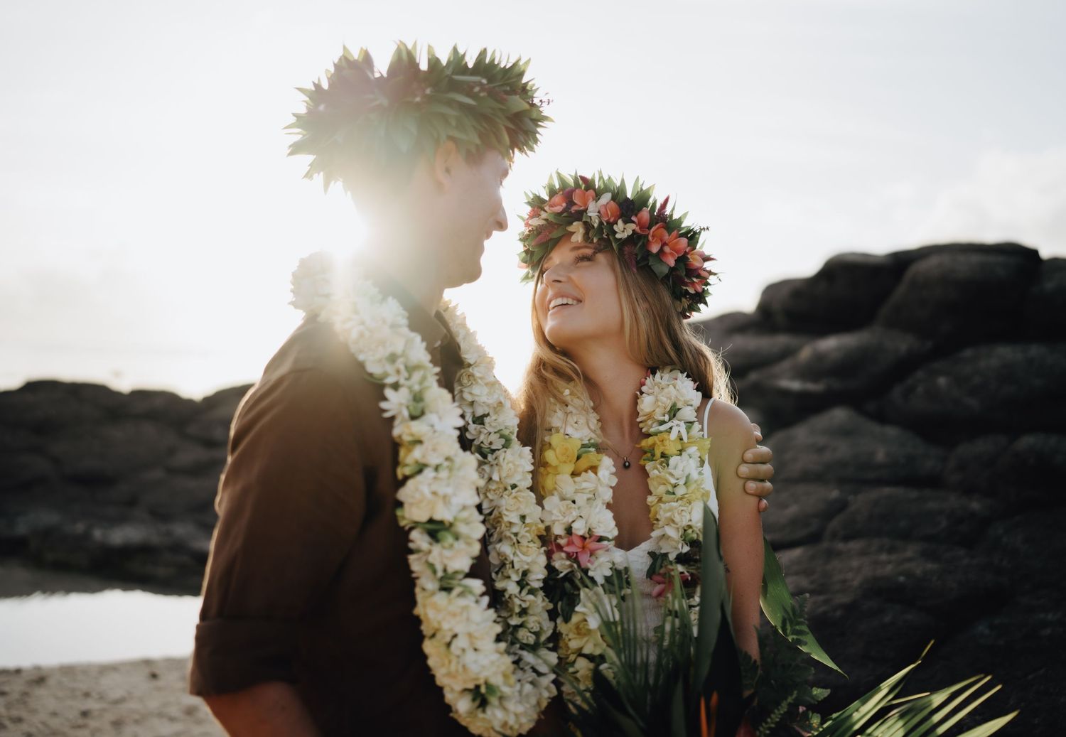 A couple in traditional Hawaiian wedding attire stands on a black sand beach at sunset with white flower leis.