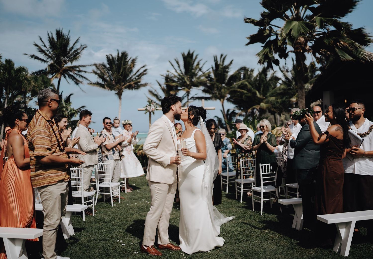 Guests celebrate at an outdoor wedding ceremony with white chairs and palm trees along a scenic coastline.