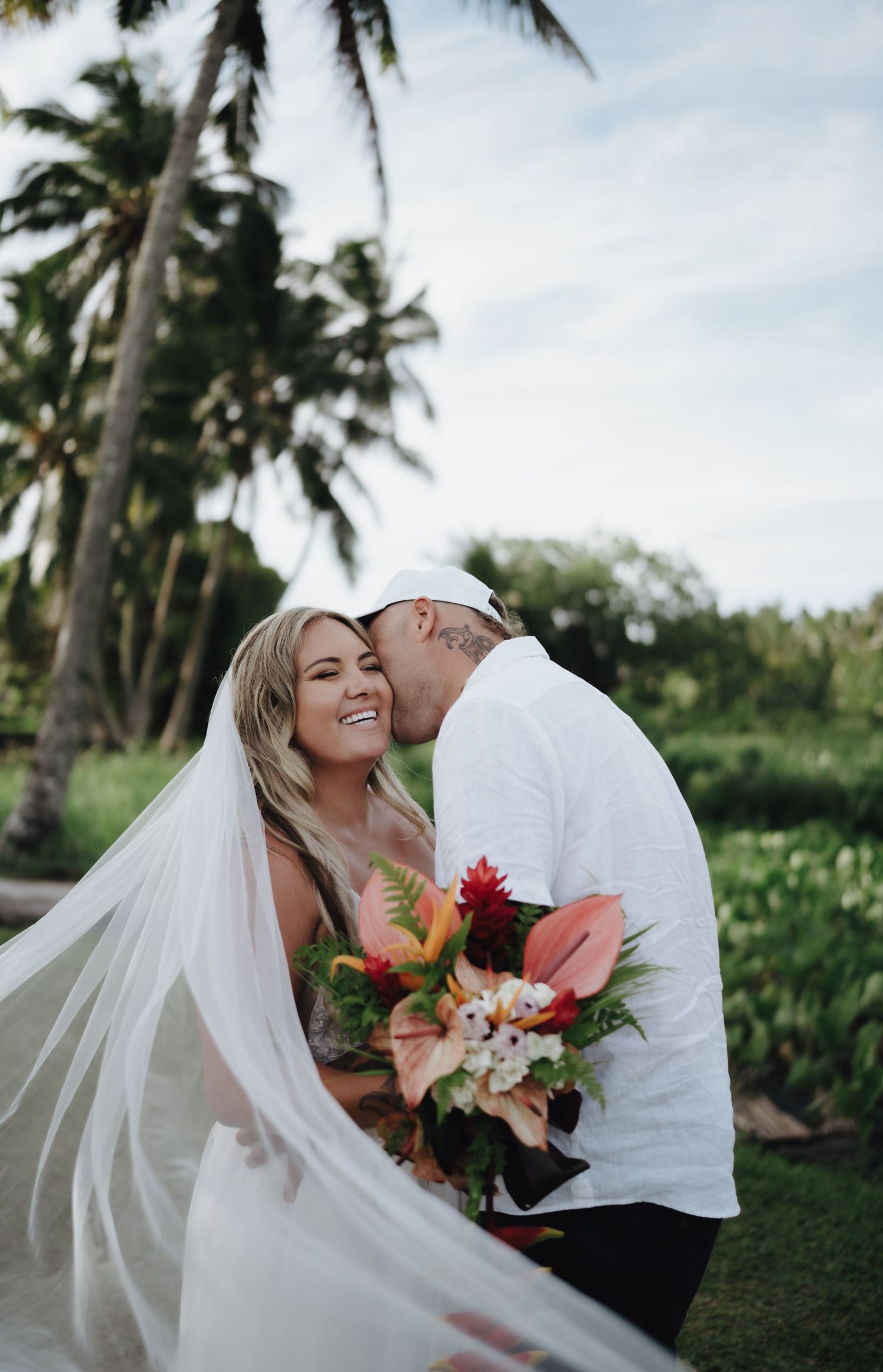 Bride in white gown and veil holds tropical bouquet as groom kisses her cheek among palm trees.