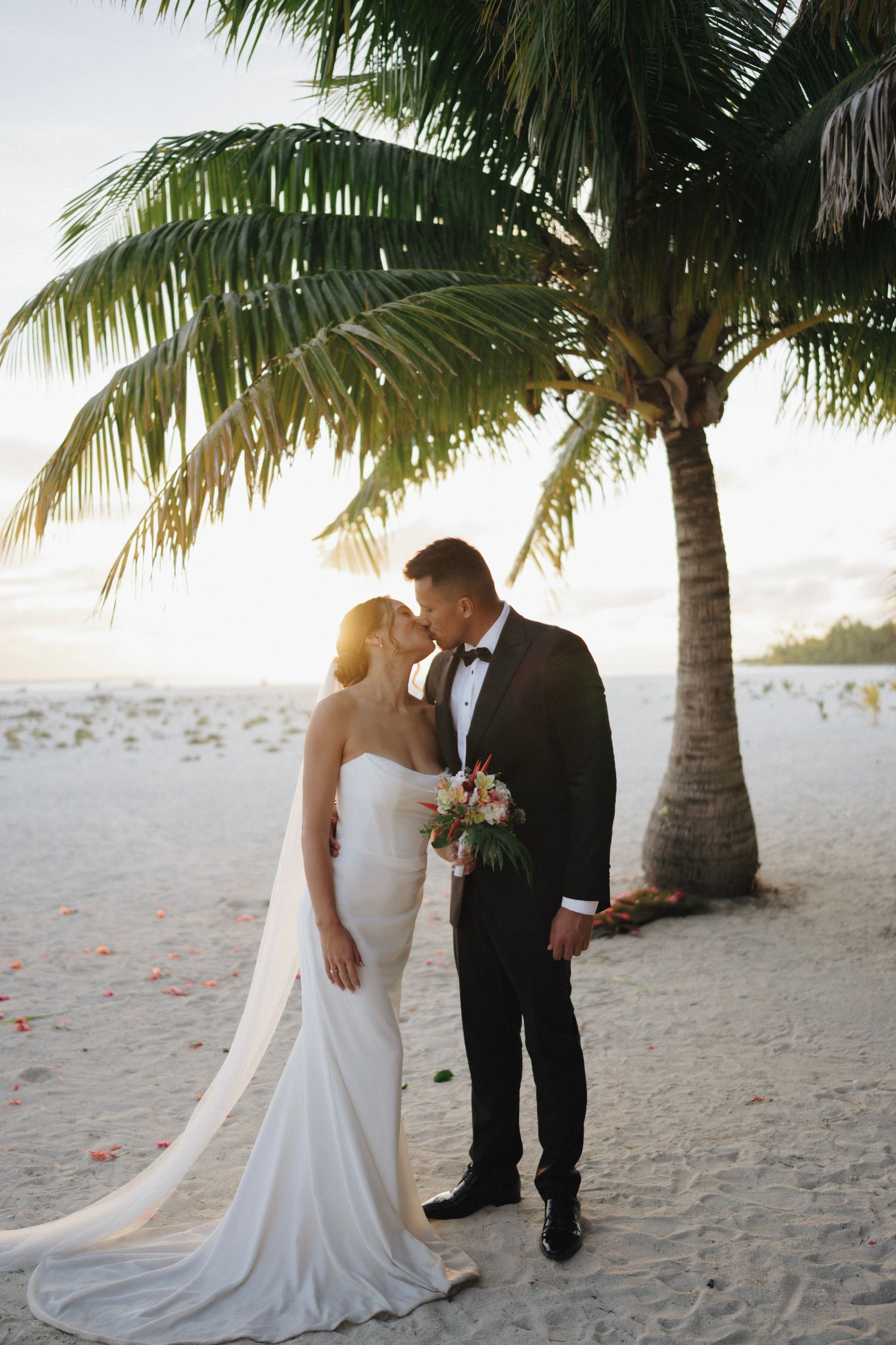 Bride and groom share a romantic kiss under a palm tree on a sandy beach at sunset, bride in white gown, groom in tuxedo.