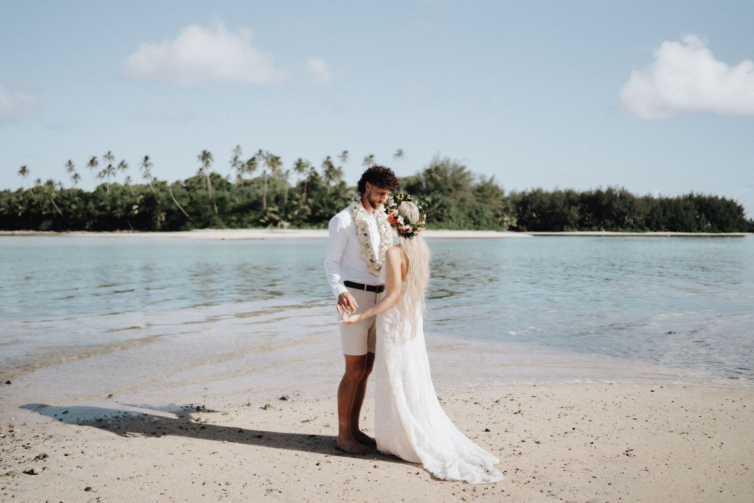 Couple sharing a romantic kiss on a tropical beach in wedding attire, with turquoise water and palm trees behind them.
