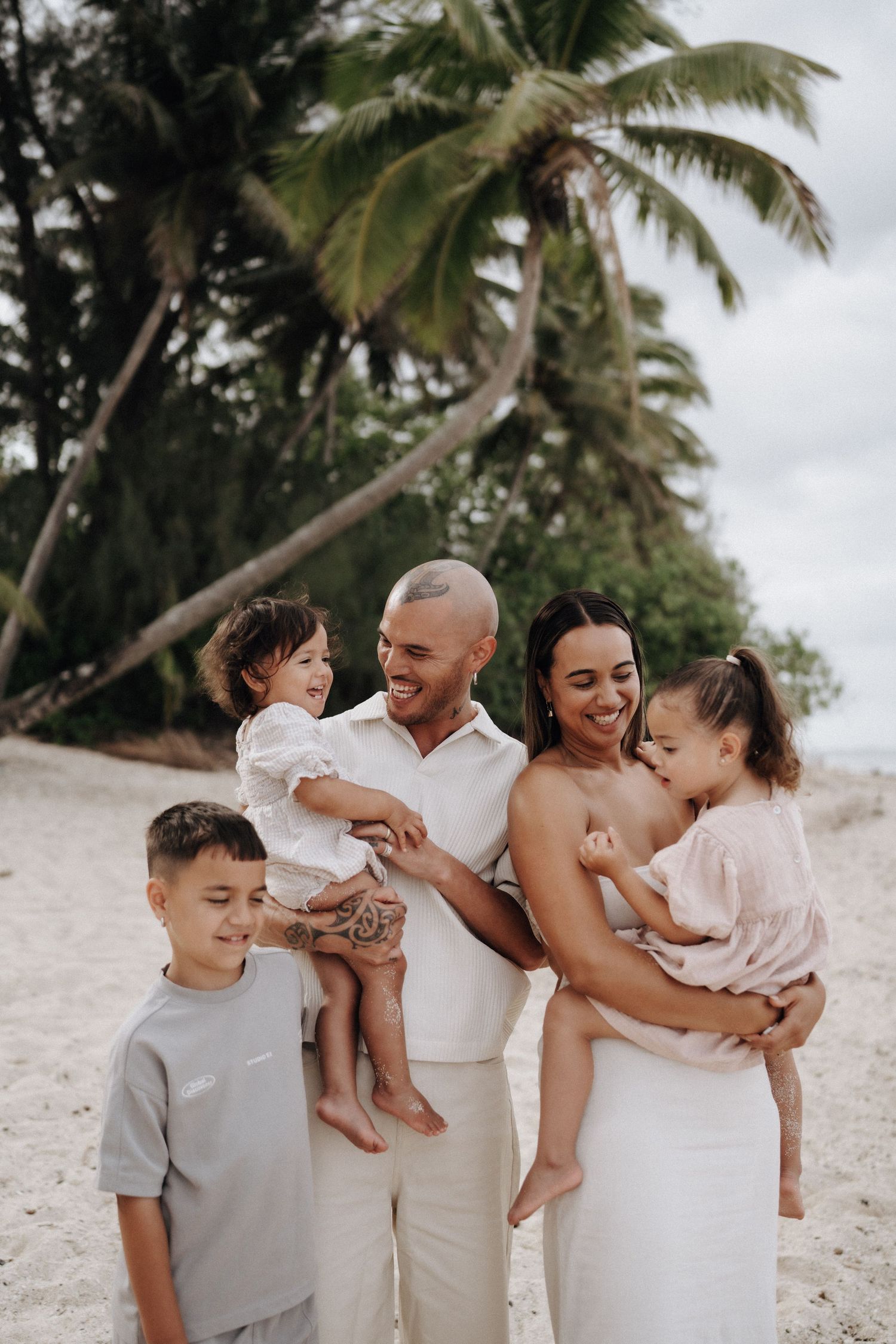 Happy family of five smiling on a tropical beach with palm trees, parents holding young children in casual white outfits.