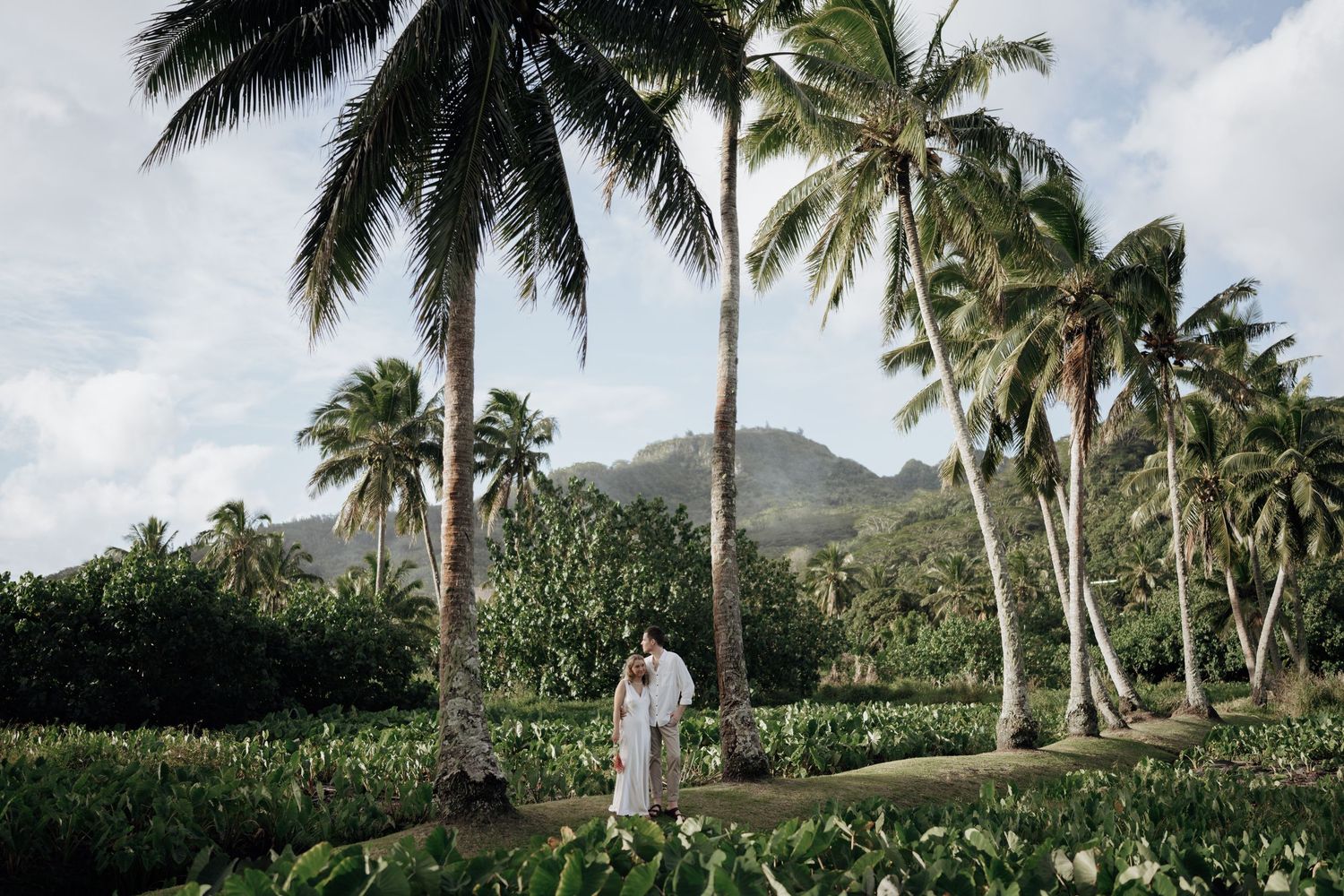 Couple in white attire standing among tropical palm trees and lush greenery with a mountain backdrop.