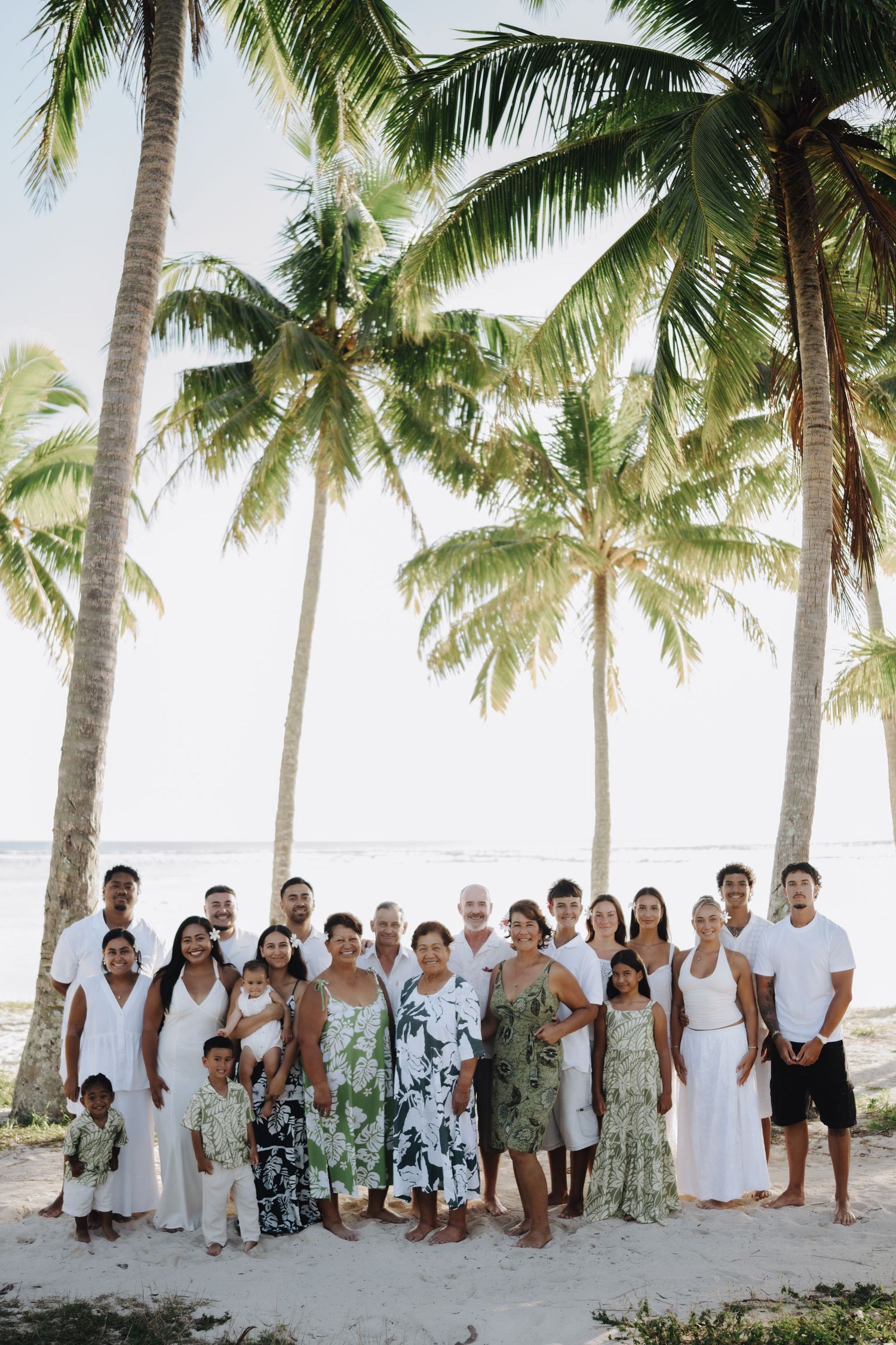 Large group in coordinated white and tropical print outfits posing together on a palm tree-lined beach.