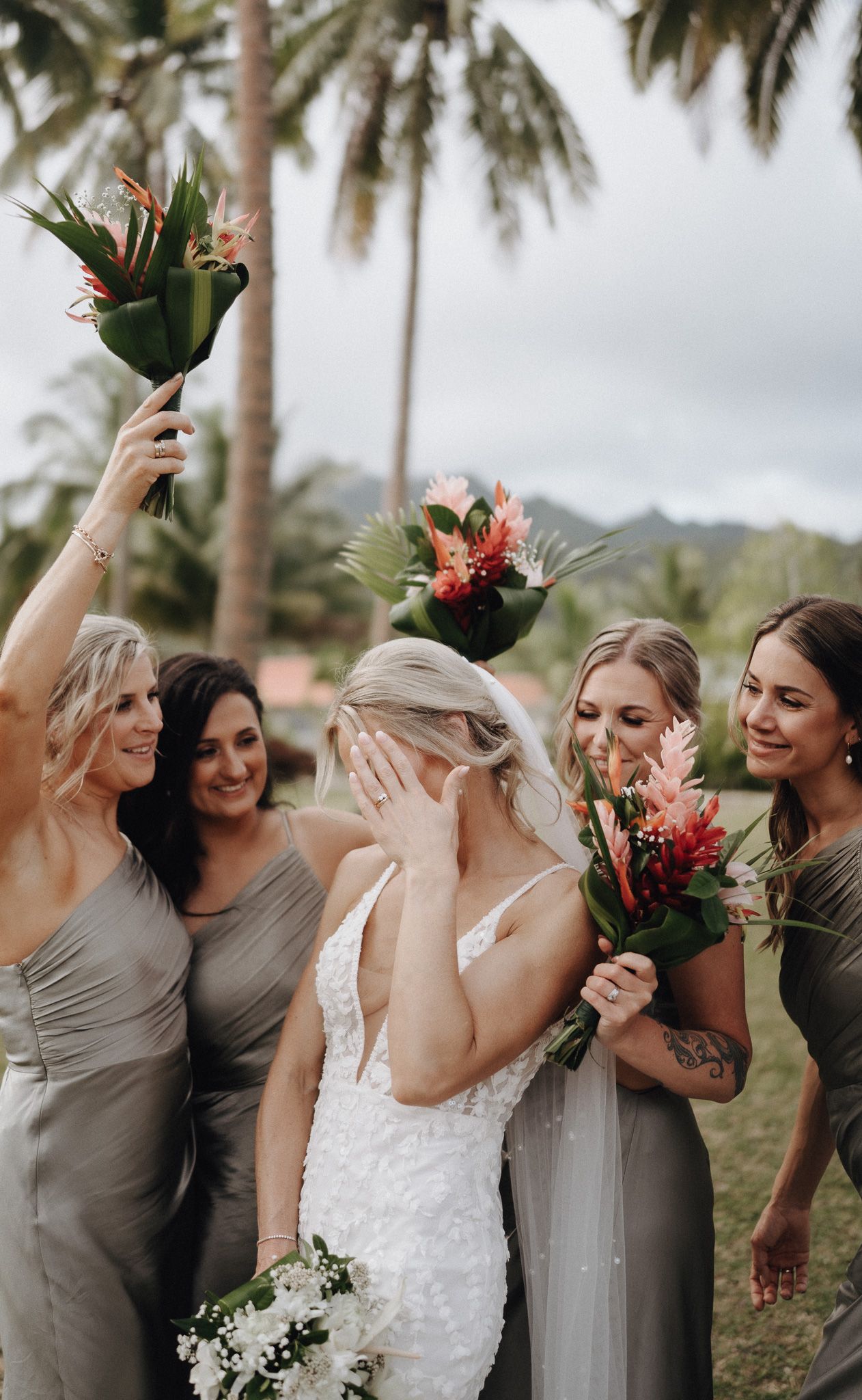 Bridesmaids in neutral dresses share a joyful moment with tropical palm trees and colorful bouquets in Hawaii.