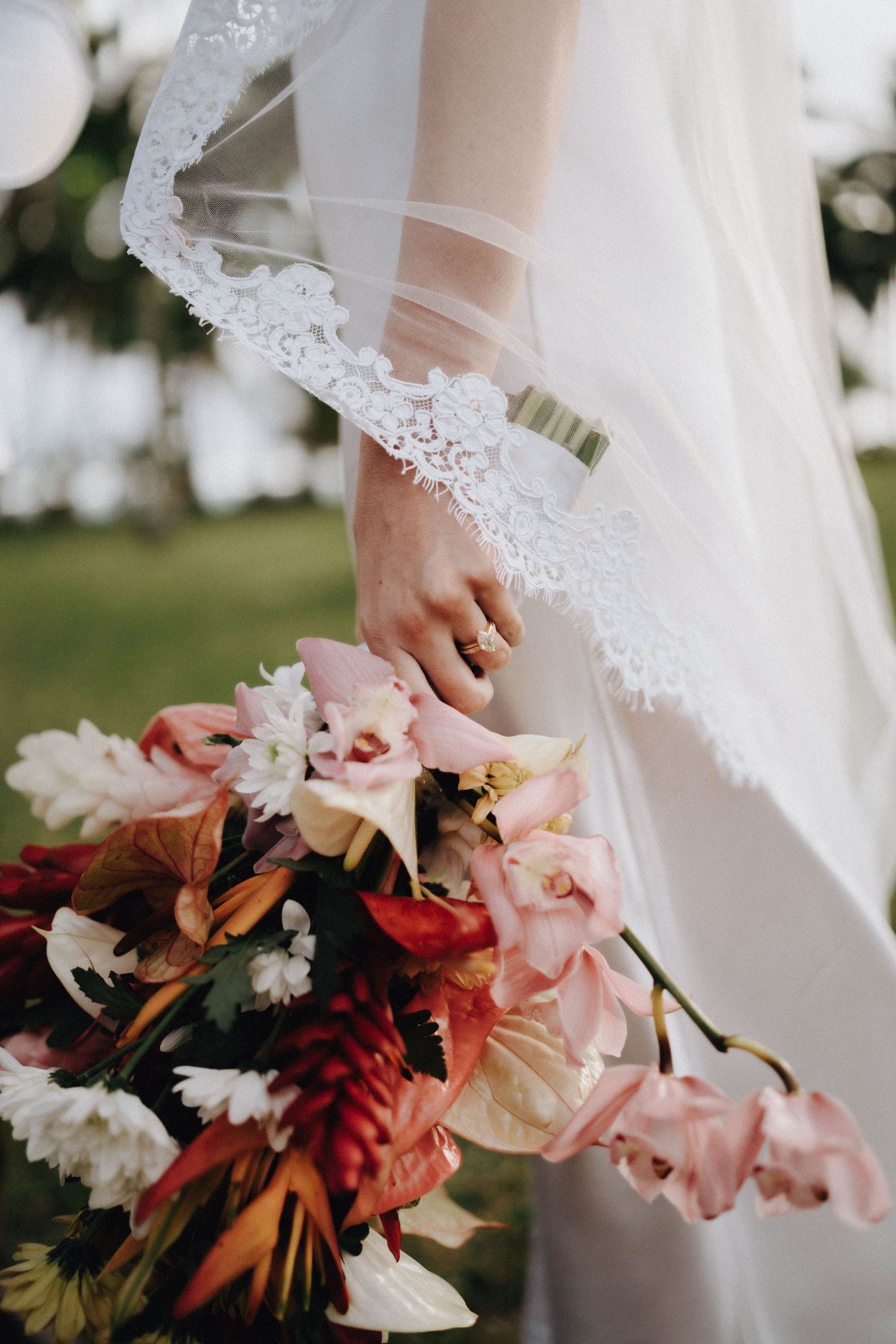 A delicate bridal bouquet with pink and burgundy flowers is held against a white lace wedding dress in natural lighting.