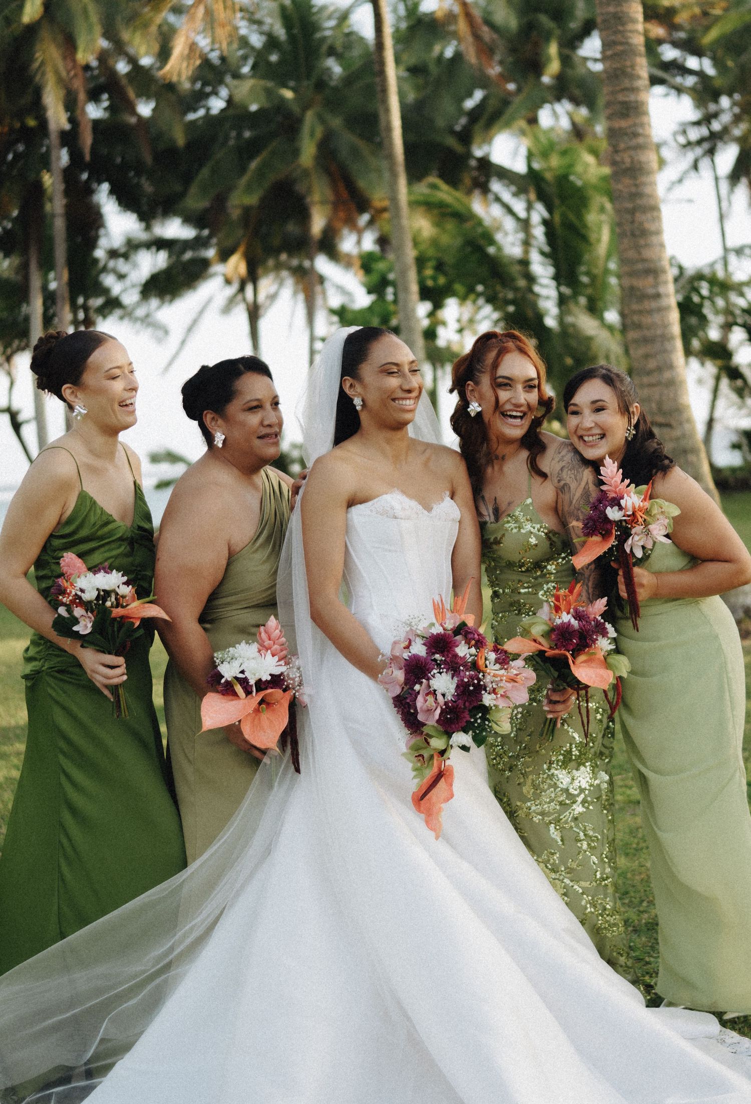 Bride in white gown with four bridesmaids in green dresses holding tropical bouquets, laughing outdoors near palm trees.