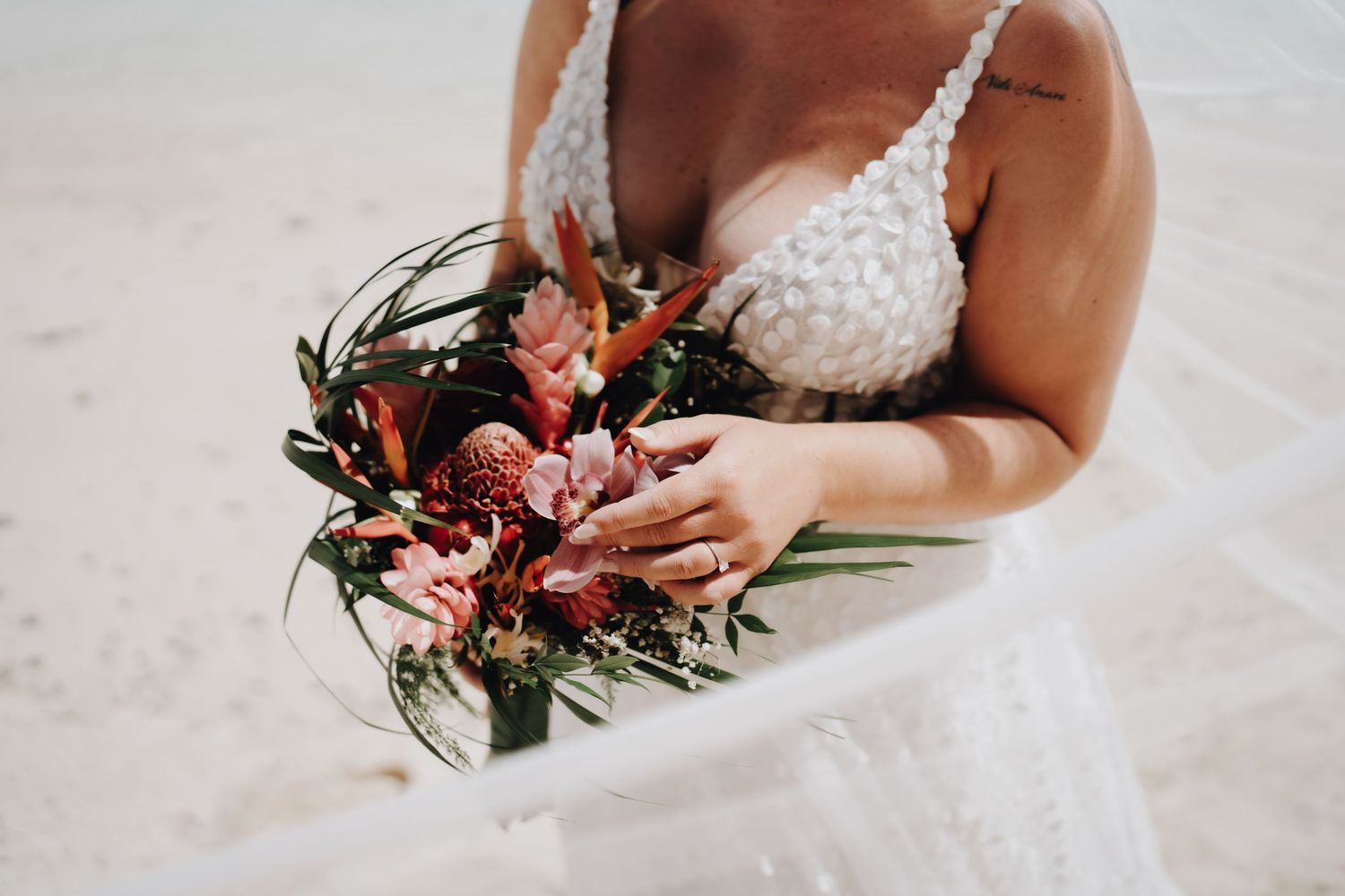 A person in a white wedding dress holds a vibrant red and green floral bouquet against a light background.