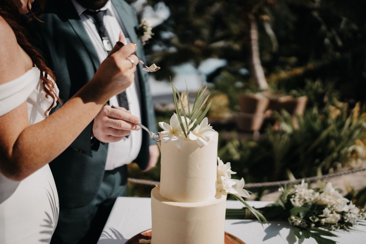Wedding guests gathered around an elegant white tiered cake topped with delicate floral decorations during an outdoor reception.