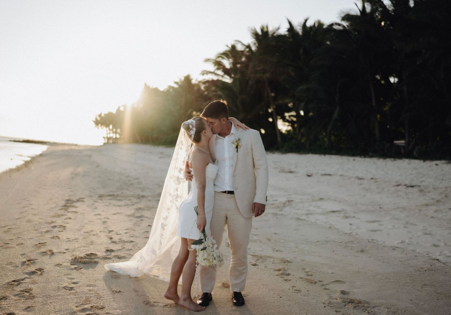 Two people in light-colored attire share an intimate moment on a beach at sunset with palm trees in the background.