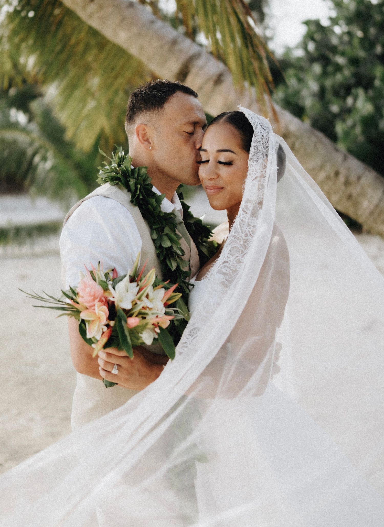 Bride and groom share a romantic kiss on a tropical beach, with a flowing veil and floral bouquet.