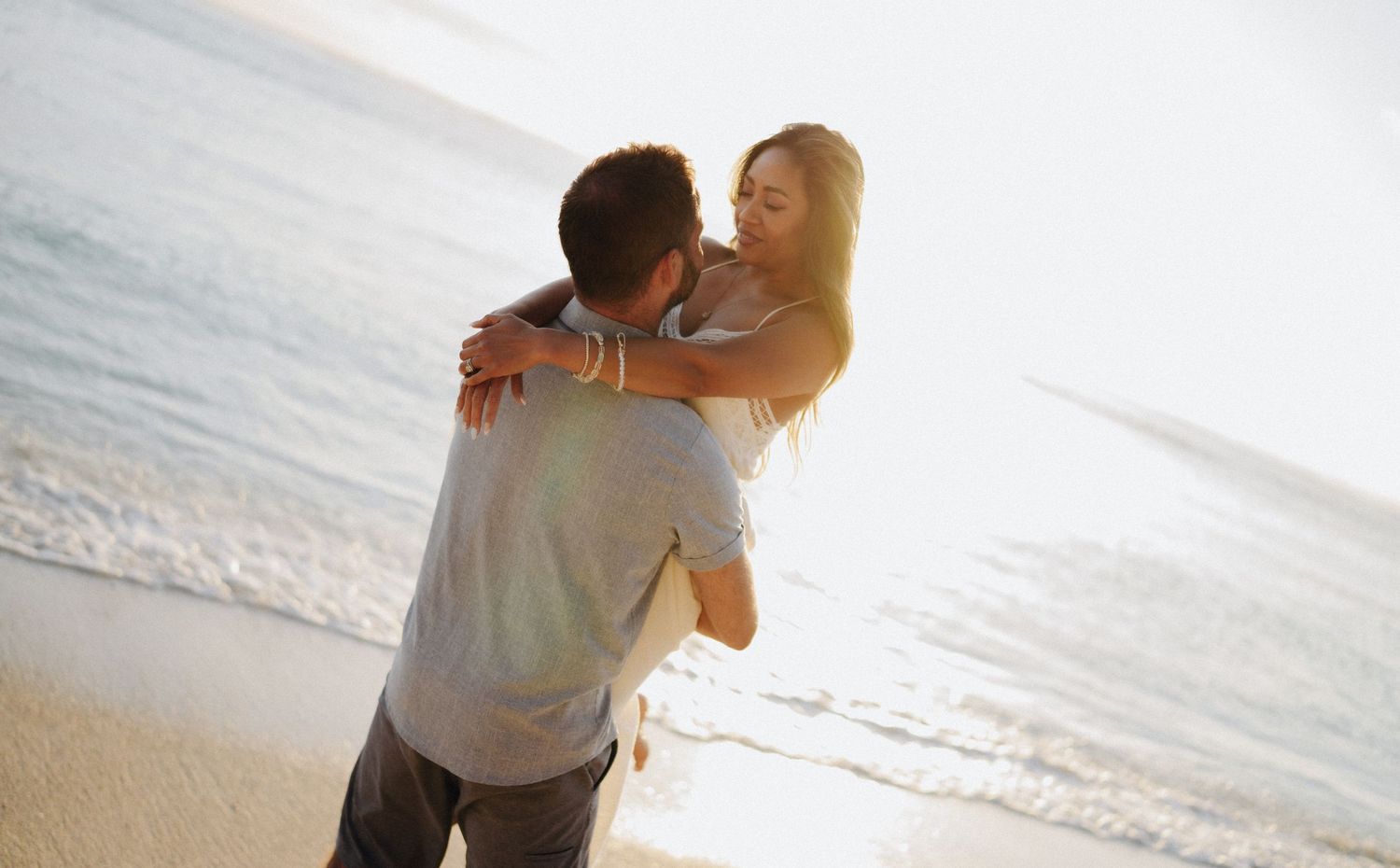 Couple embracing on a sunny beach, woman lifted in man's arms with ocean waves in the background.