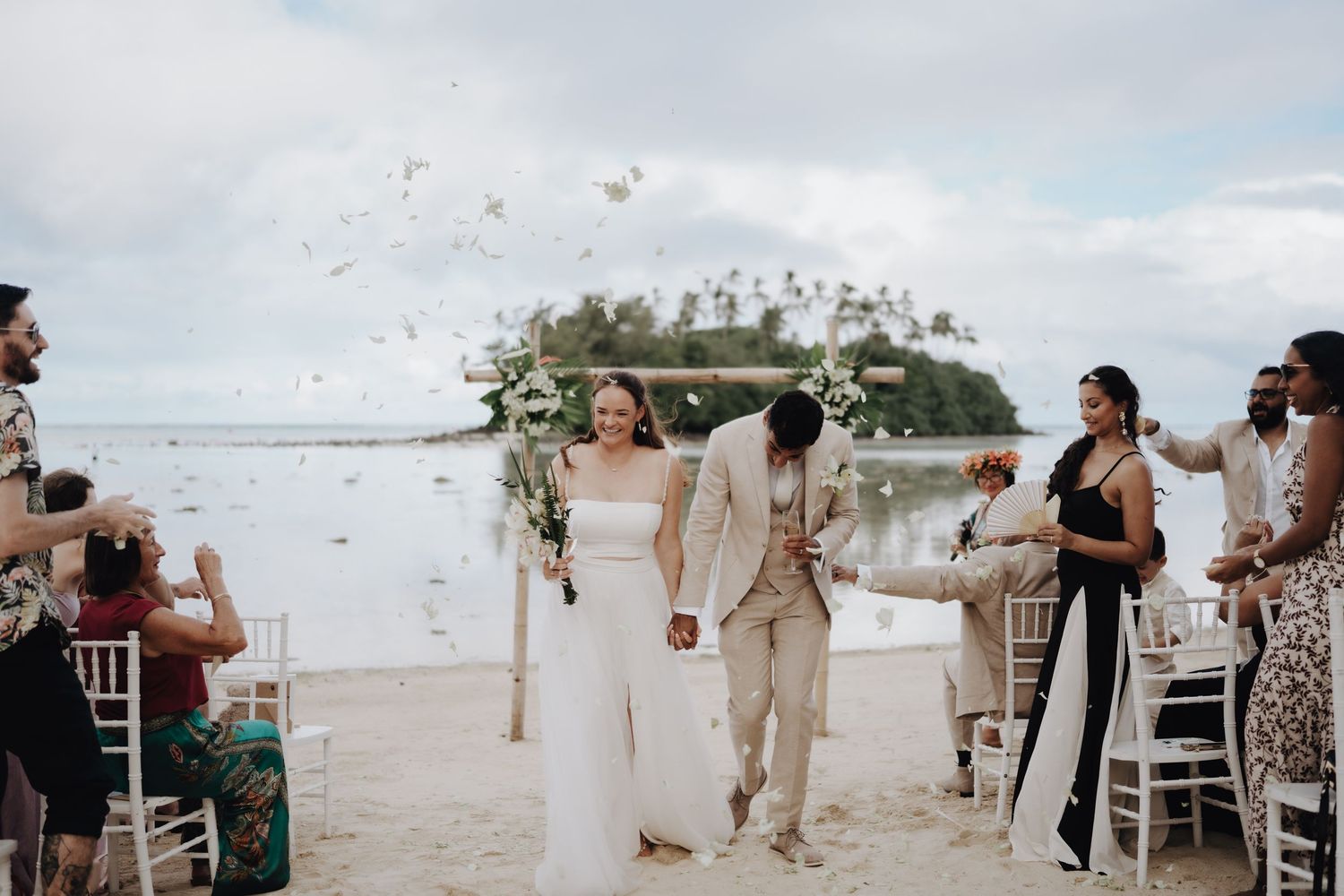 Guests celebrate at a beachside wedding reception with tables and chairs set up on white sand near the ocean shore.