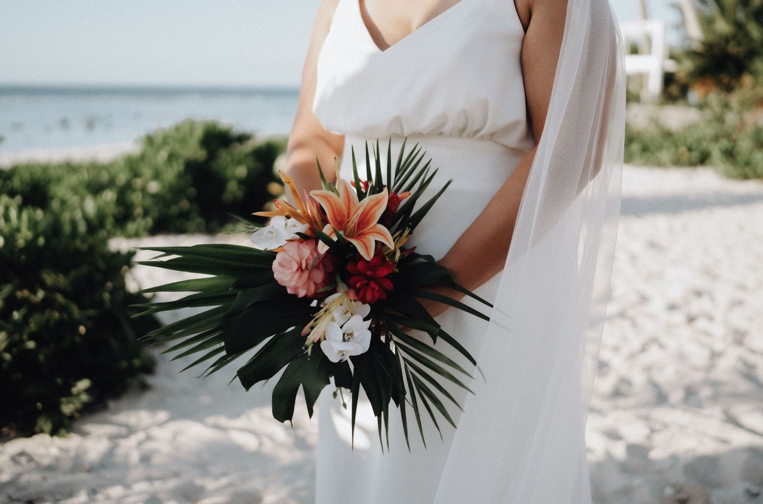 A beach wedding scene with a white dress and tropical bouquet featuring lilies and palm fronds against sandy backdrop.