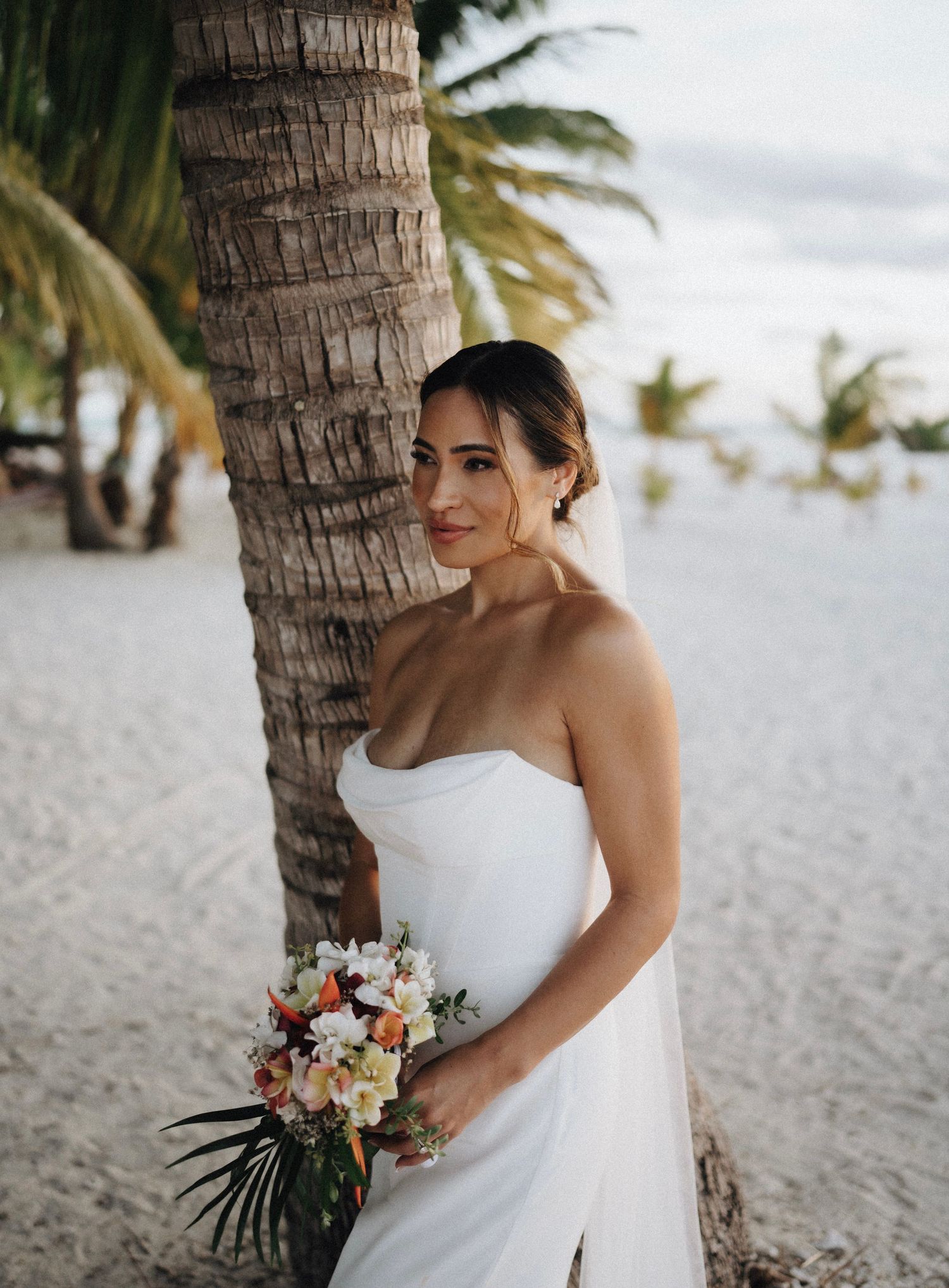 Bride in strapless white gown holding tropical bouquet leaning against palm tree on sandy beach.