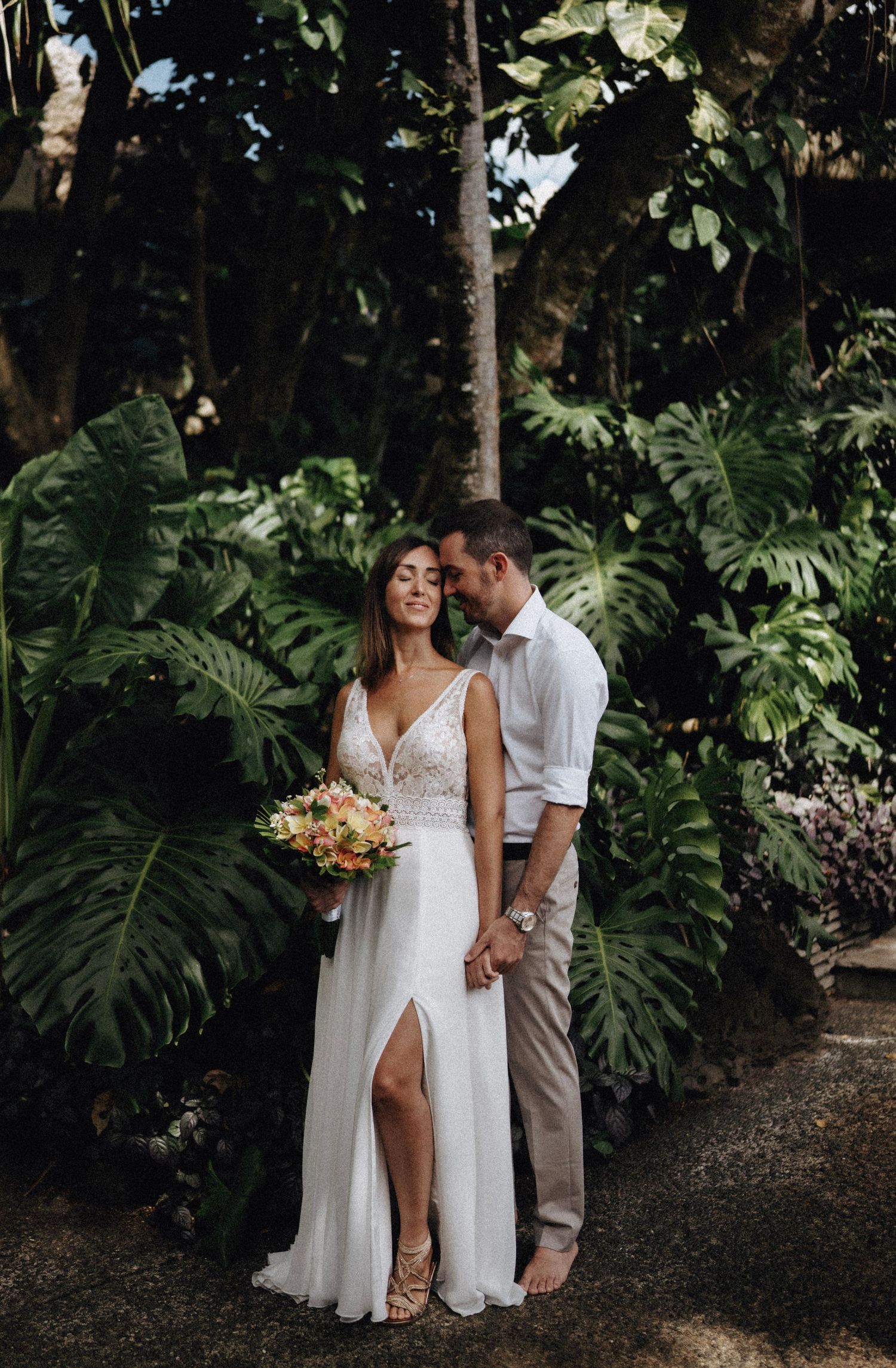 A couple shares an intimate moment in a tropical garden setting with lush monstera leaves and white flowing dress.