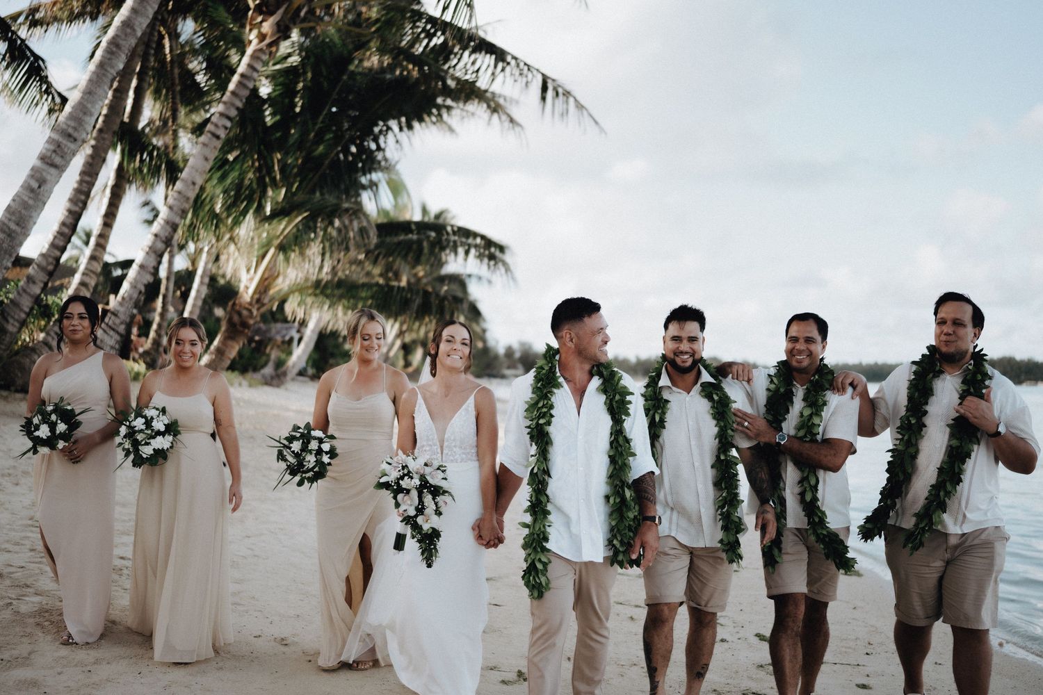 Wedding party walks along a tropical beach with palm trees, wearing beige attire and traditional Hawaiian leis.