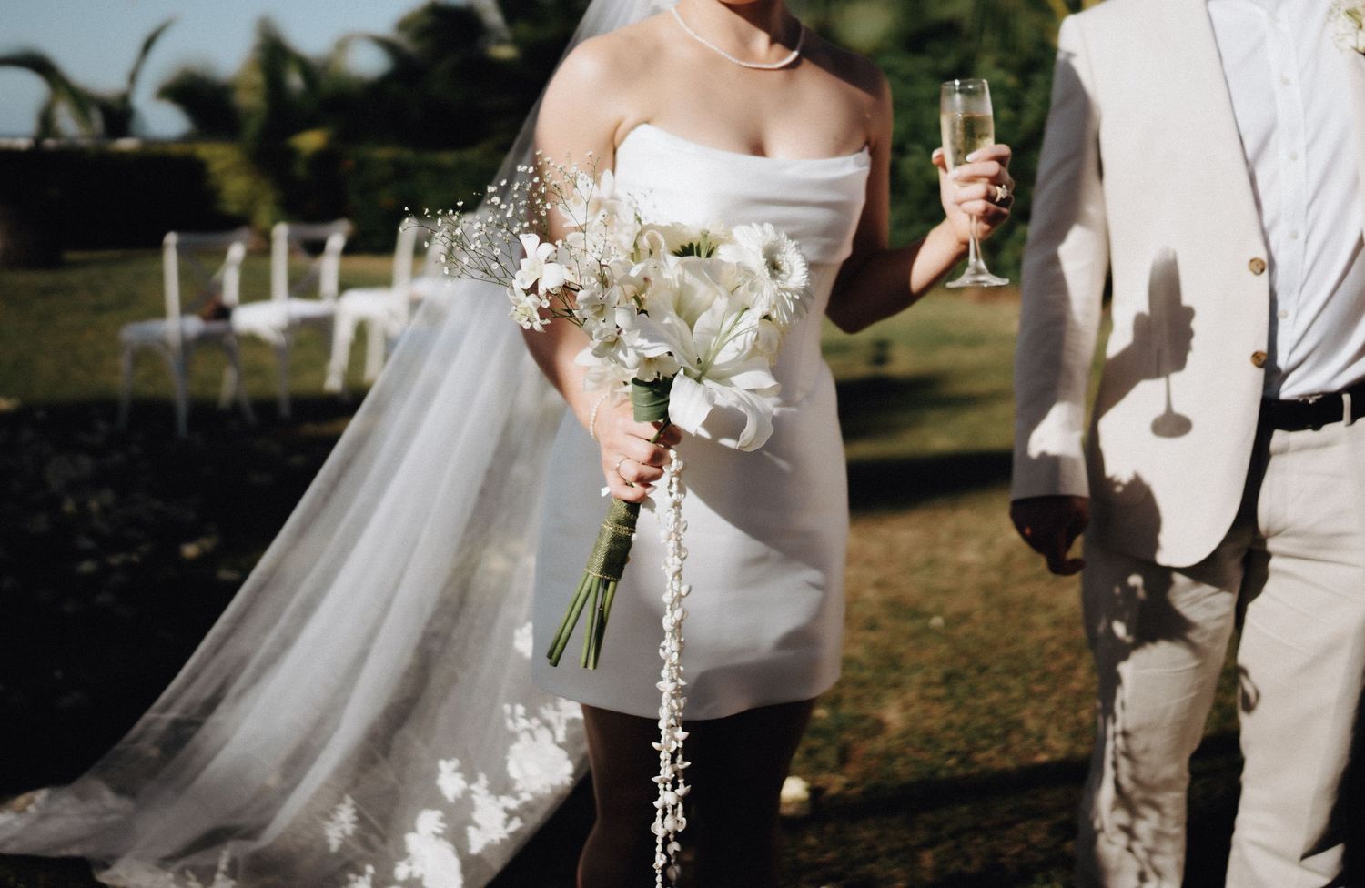 A couple shares a celebratory moment with champagne glasses while holding a delicate white floral bouquet at an outdoor wedding.