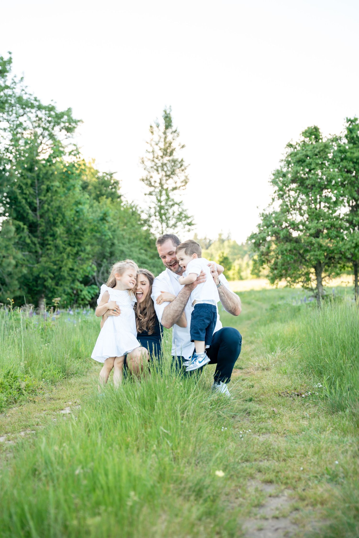 A happy family of four laughs and plays together on a grassy path surrounded by trees in summer.